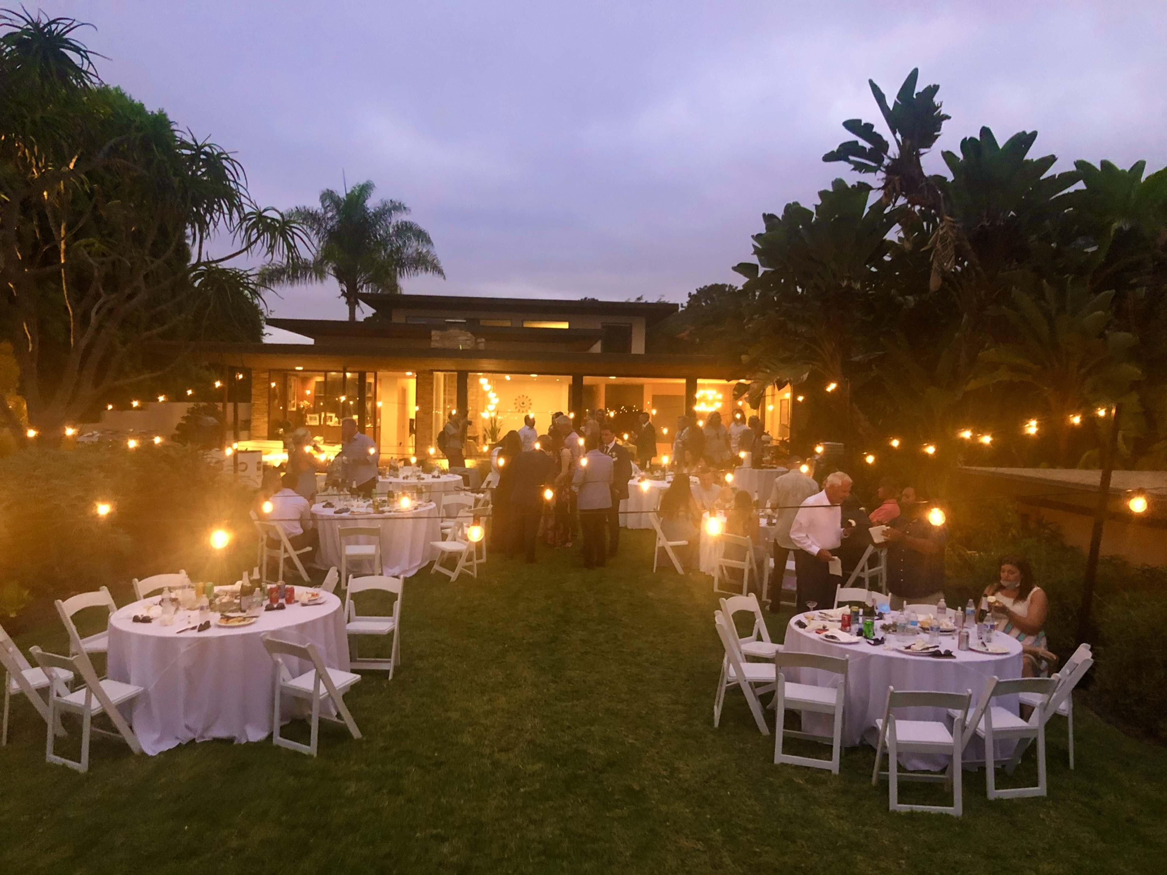 A garden party with elegantly set tables and guests mingling in soft, ambient light as night begins to fall.