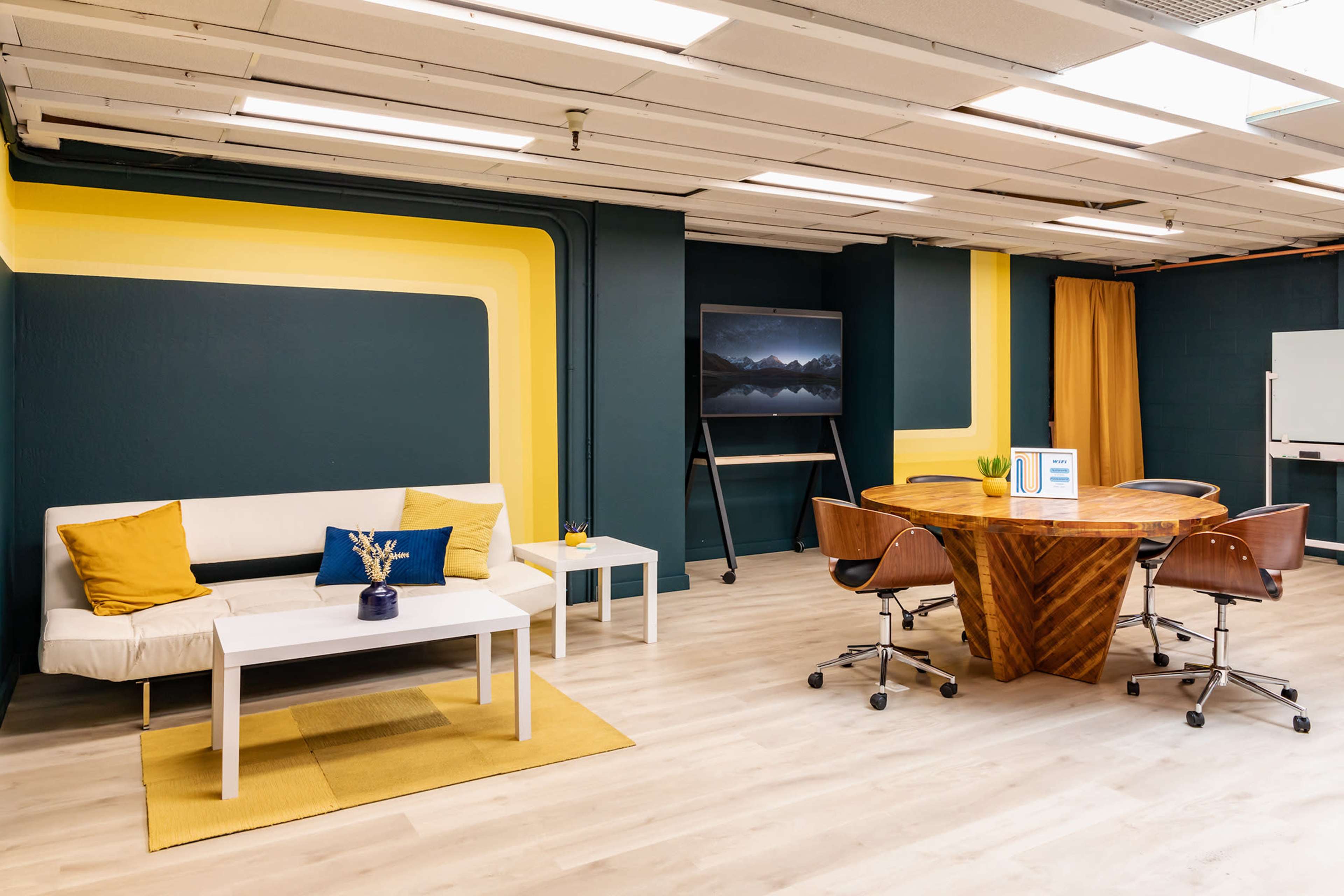 A modern office space with a white sofa, a wooden round table, and a large screen mounted on a wall, featuring blue and yellow accent colors.