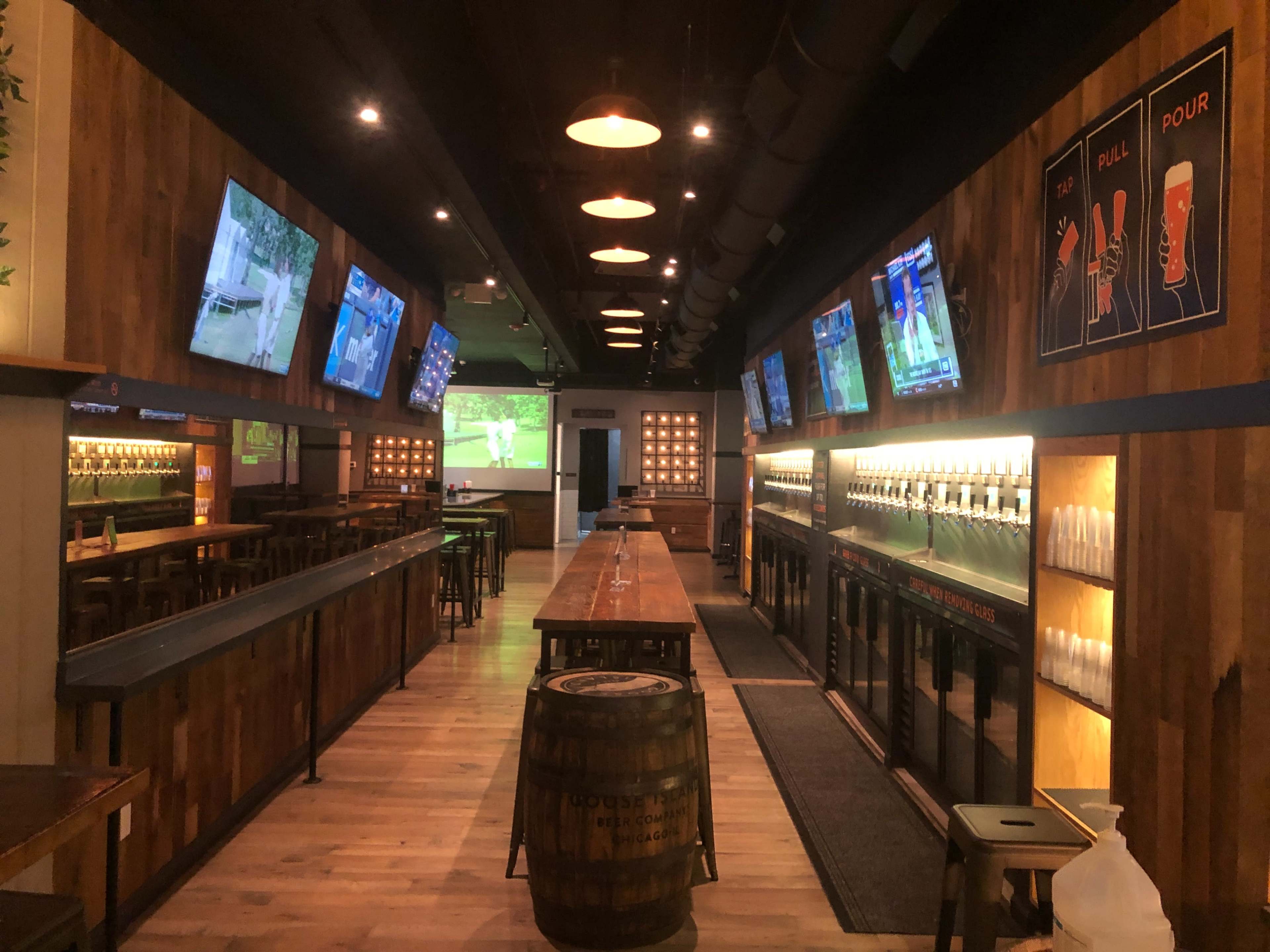 The image shows a modern bar interior with wooden walls, rows of taps, several flat-screen TVs displaying sports, and a long communal table.