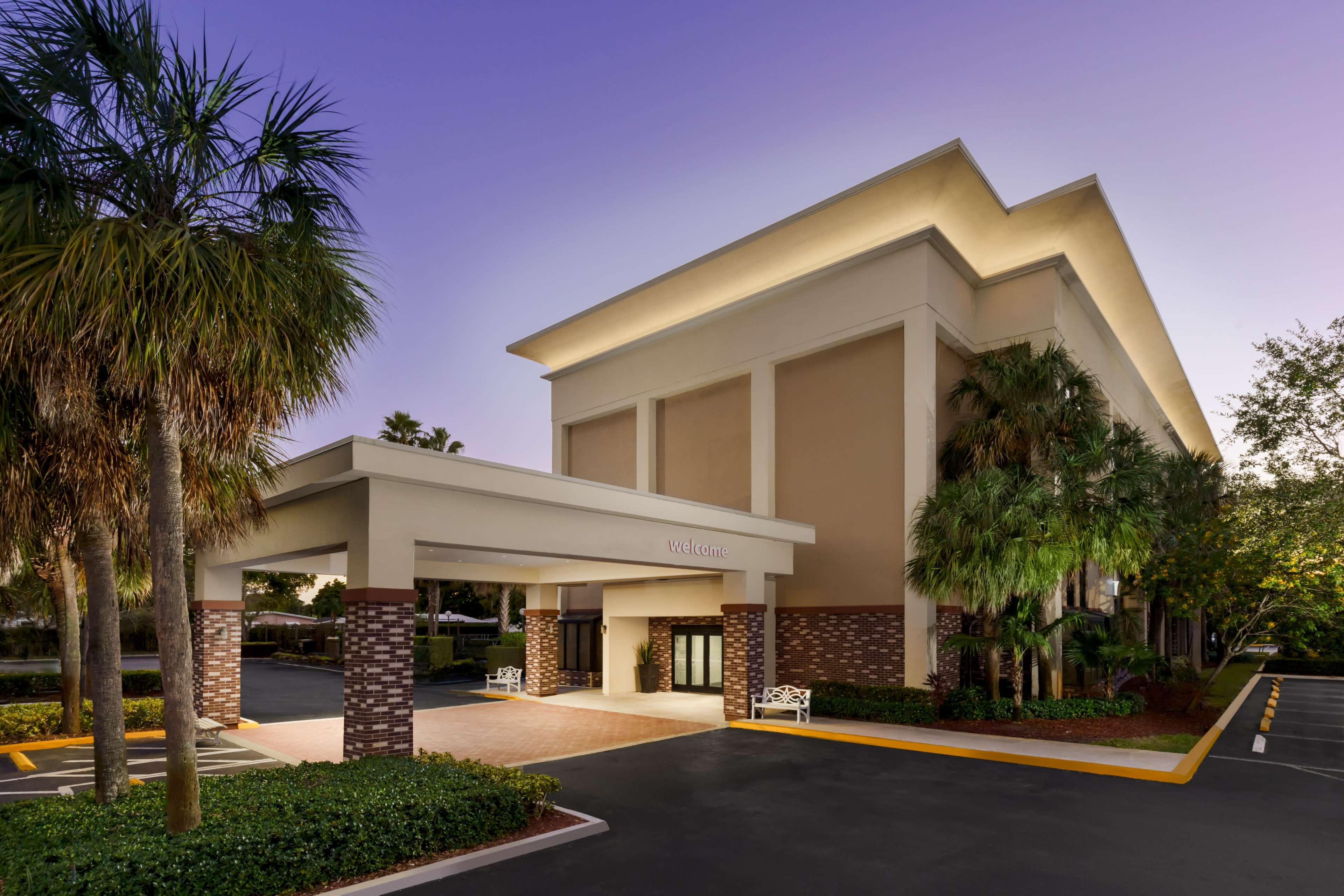 A three-story hotel with a welcoming entrance and palm trees in the vicinity is shown under a dusky sky.