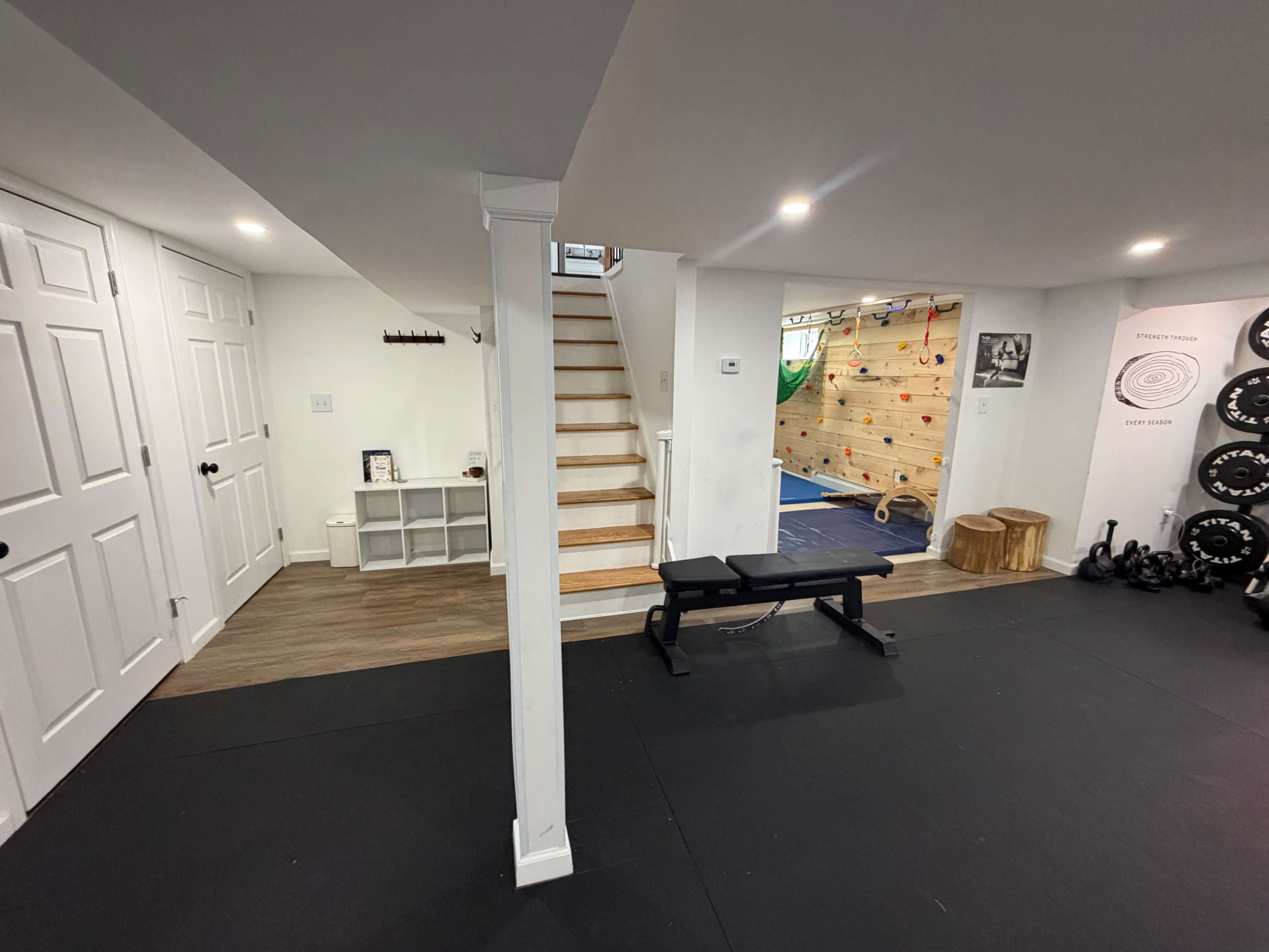The image shows a basement area featuring a workout space with exercise equipment, a climbing wall, and a staircase leading to an upper level.