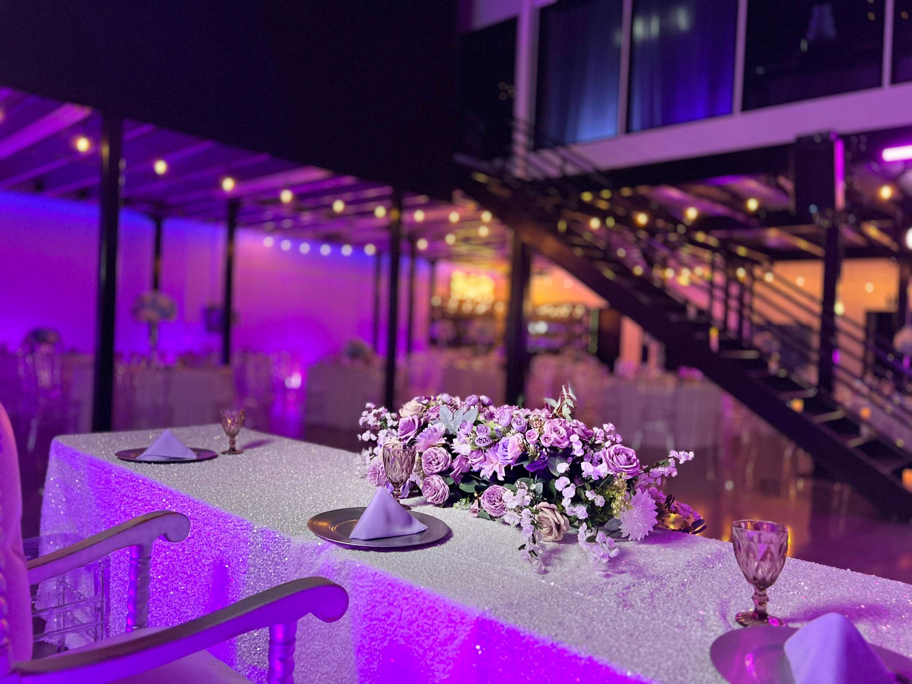 A table adorned with a floral centerpiece and elegant place settings is set in a dimly lit venue with colorful lighting and a staircase in the background.