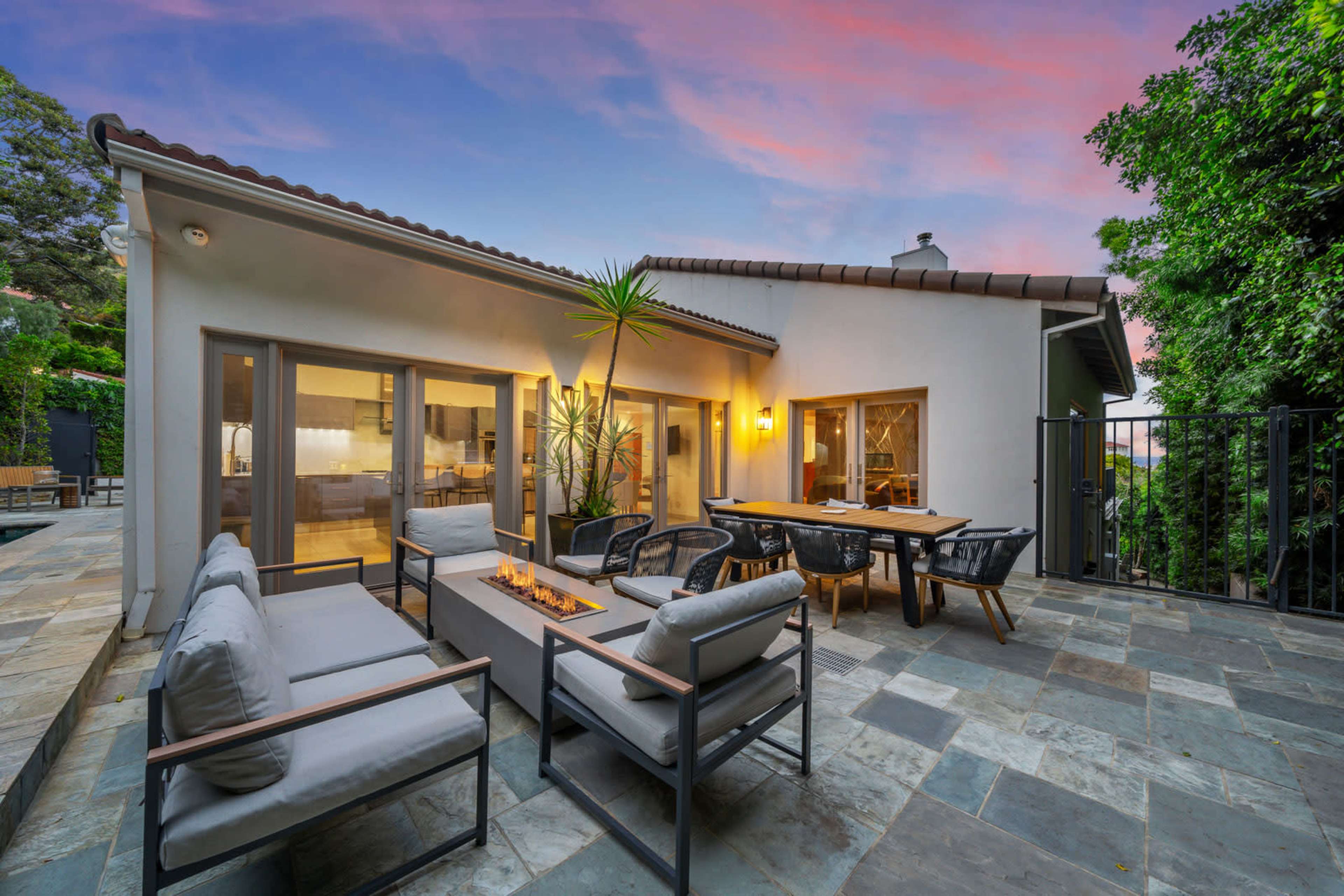 A modern patio features a seating area around a fire pit, bordered by sliding glass doors leading into a contemporary home.