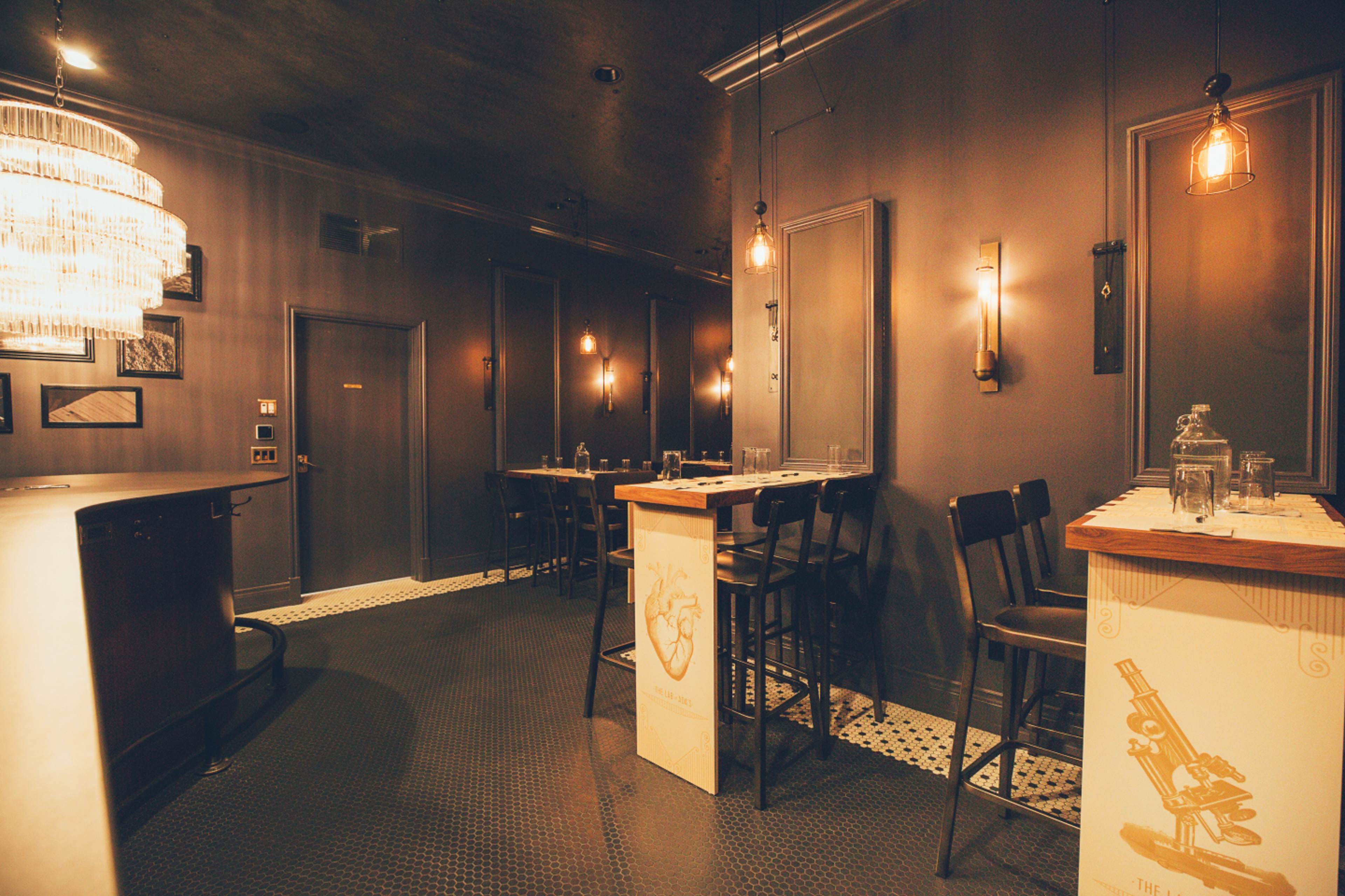A dimly lit restaurant interior featuring a bar area with high chairs, wooden tables, and a chic chandelier.