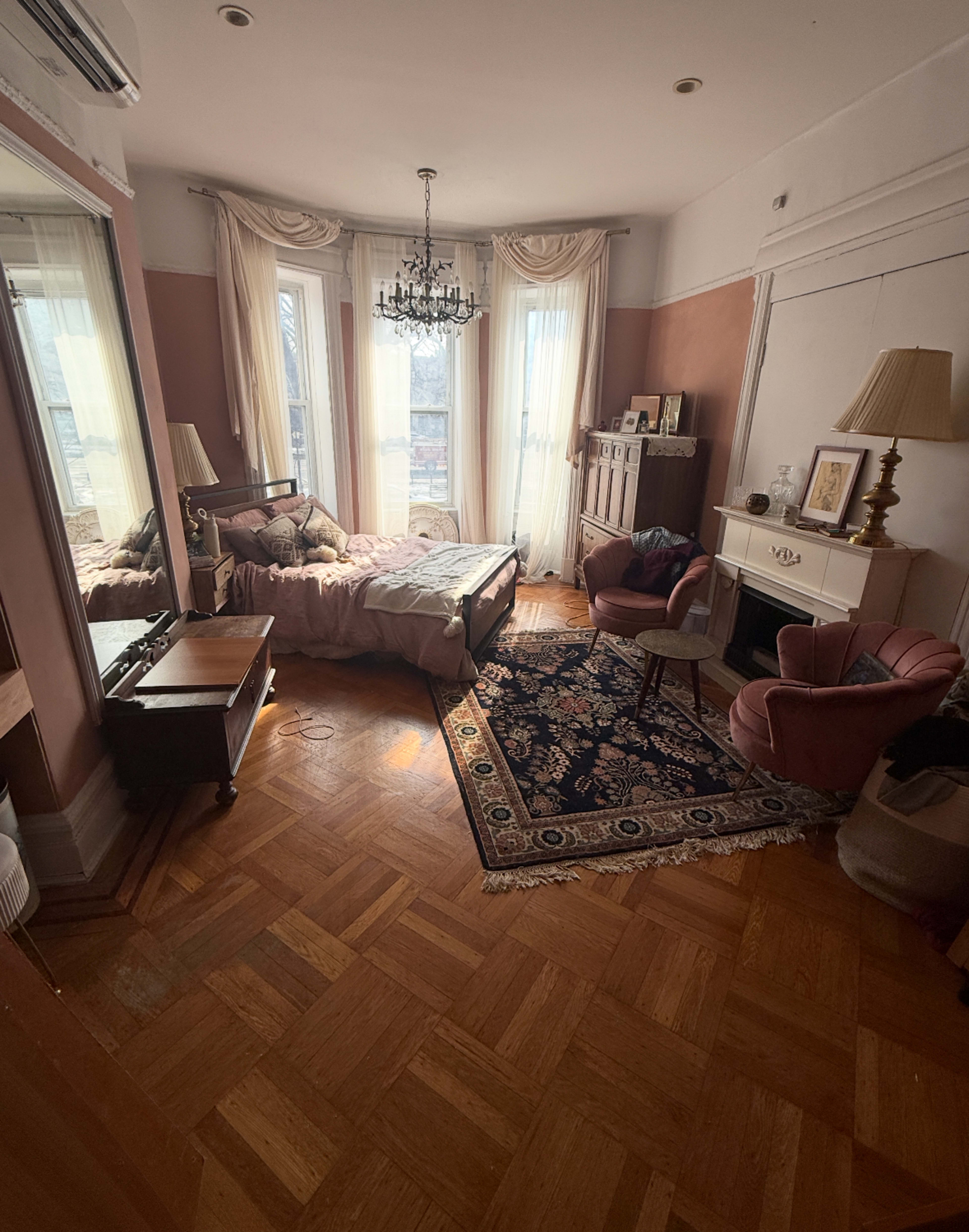 The image shows a well-furnished bedroom with a chandelier, large windows, a bed, and a decorative rug on a hardwood floor.