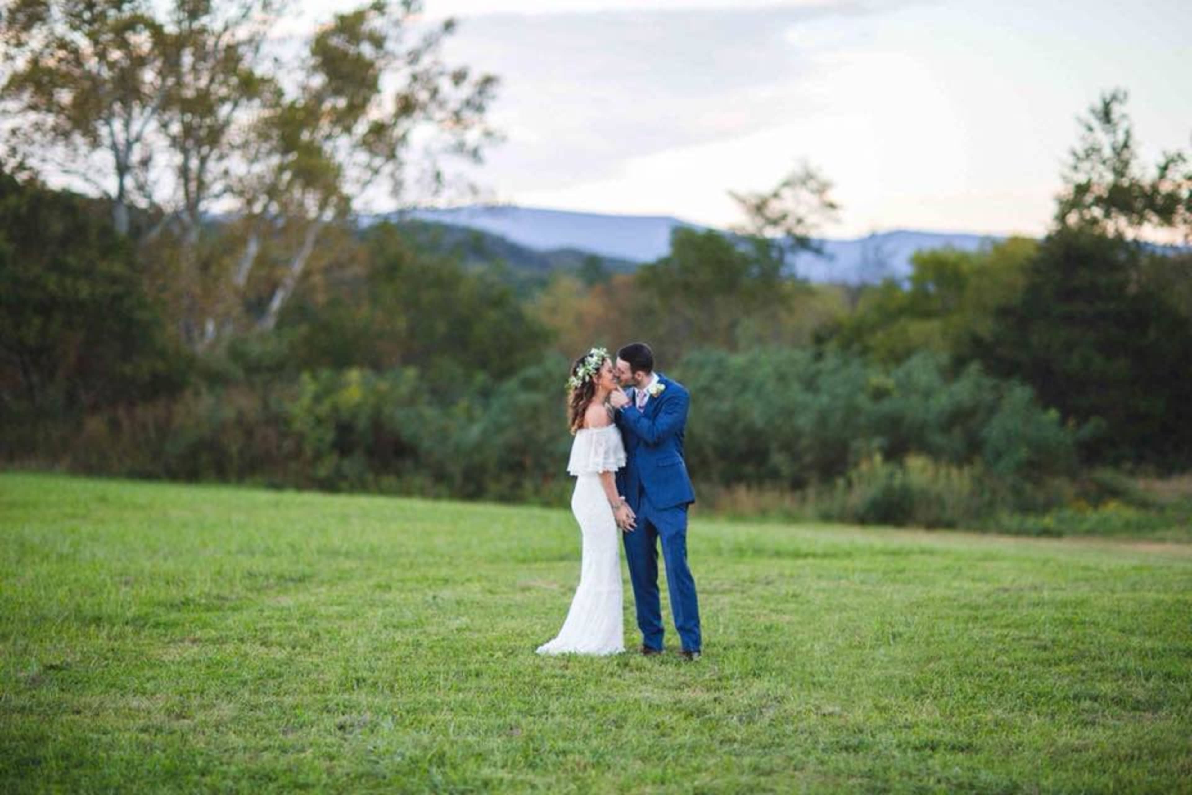 A couple stands in an open field, with trees and mountains in the background, as one leans in to embrace the other.