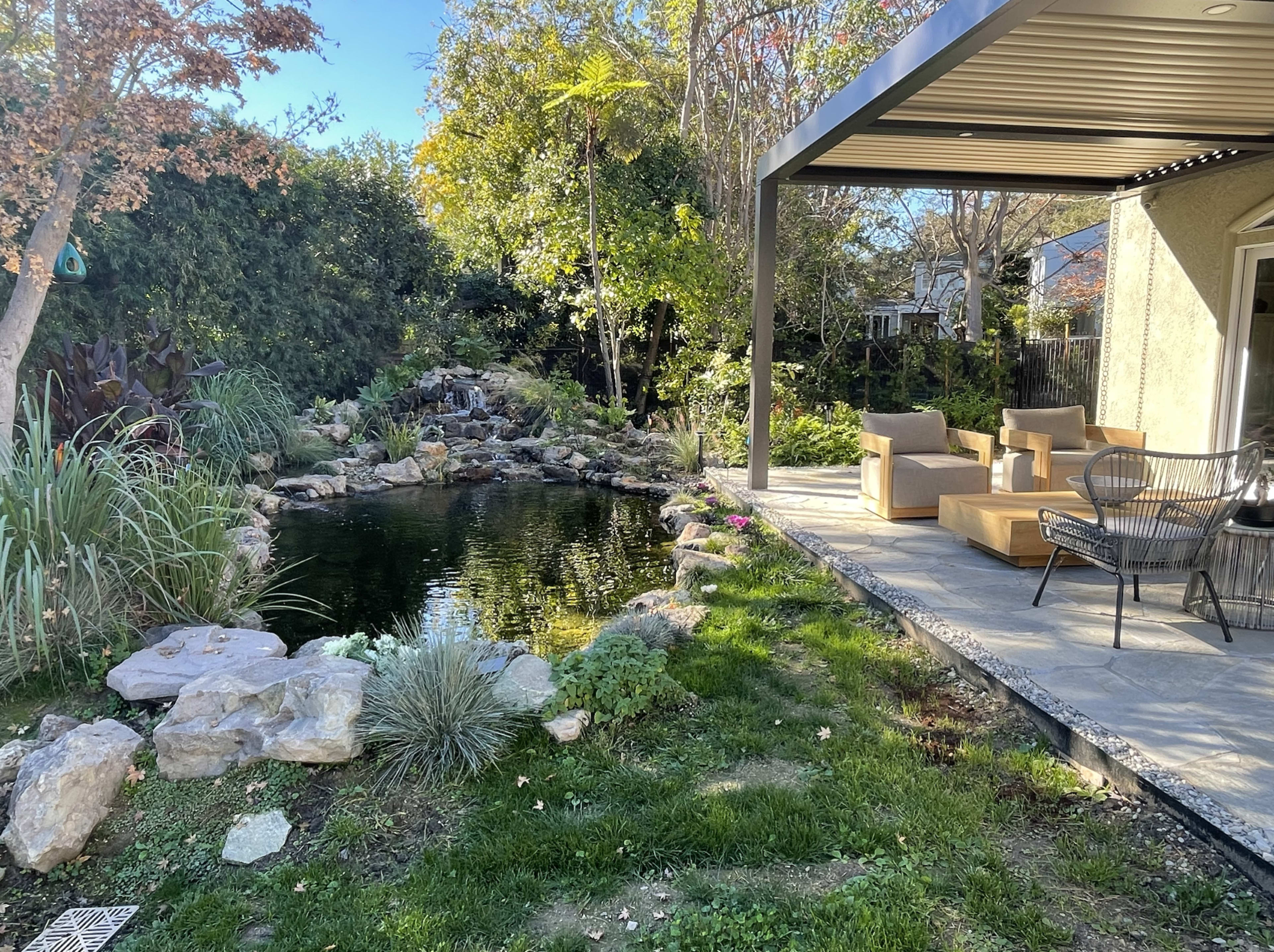 A patio area with wooden seating overlooks a pond surrounded by rocks and plants in a landscaped garden.