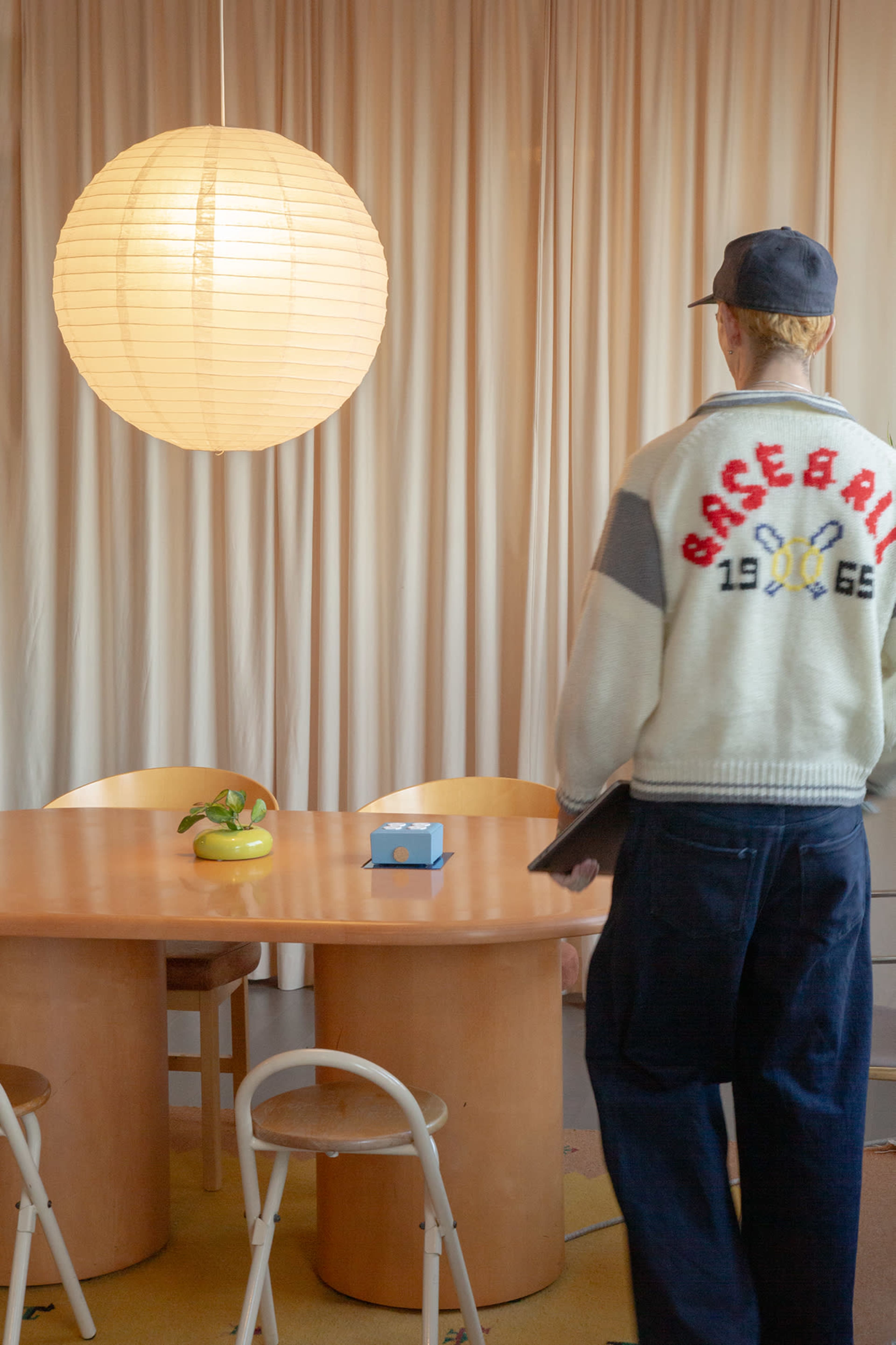 A person wearing a sweater stands beside a circular table with a vase and a small box in a softly lit room featuring a large paper lantern.