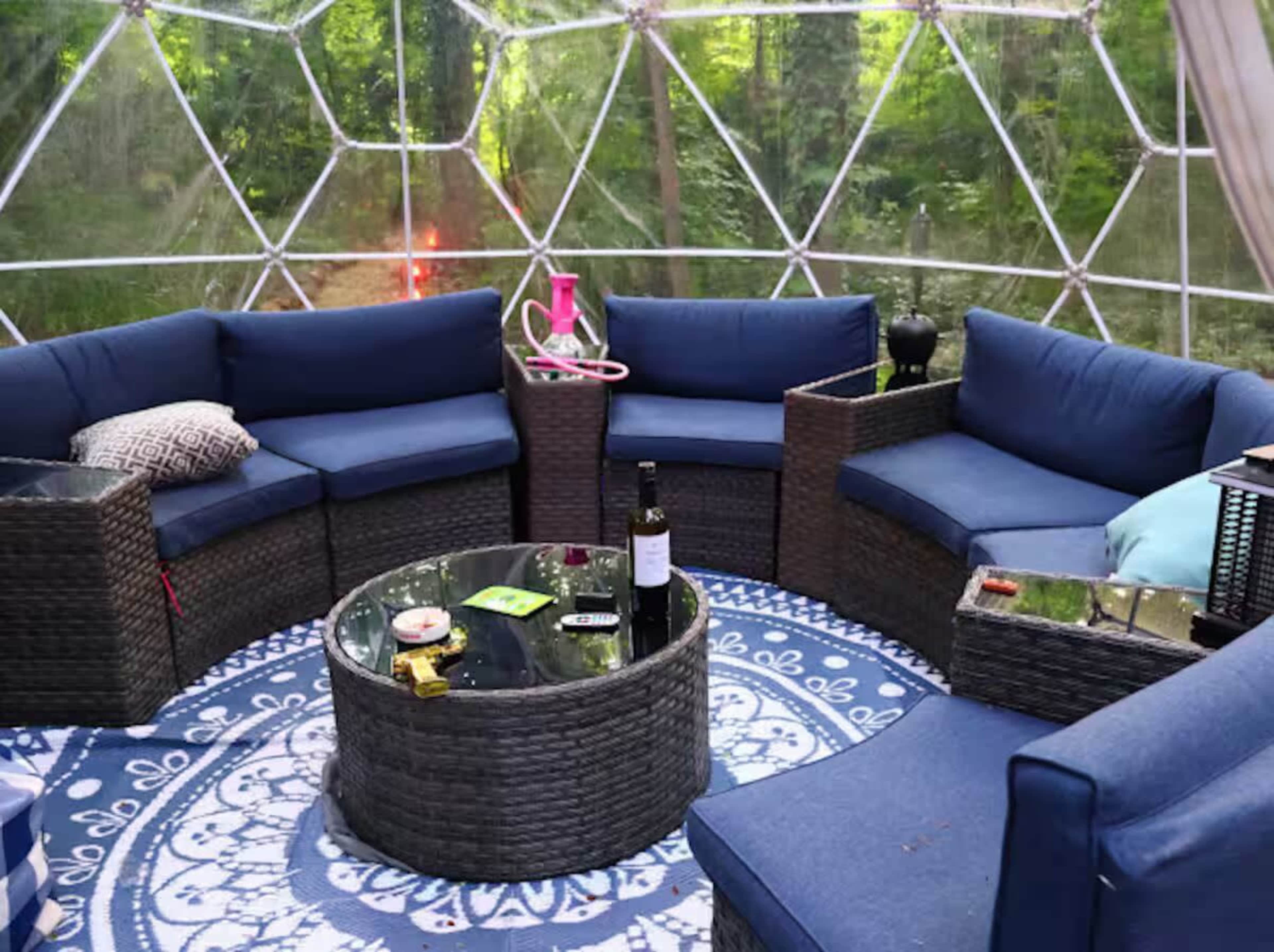 The image shows a geodesic dome interior furnished with circular seating arranged around a glass-top table, surrounded by a forest backdrop.