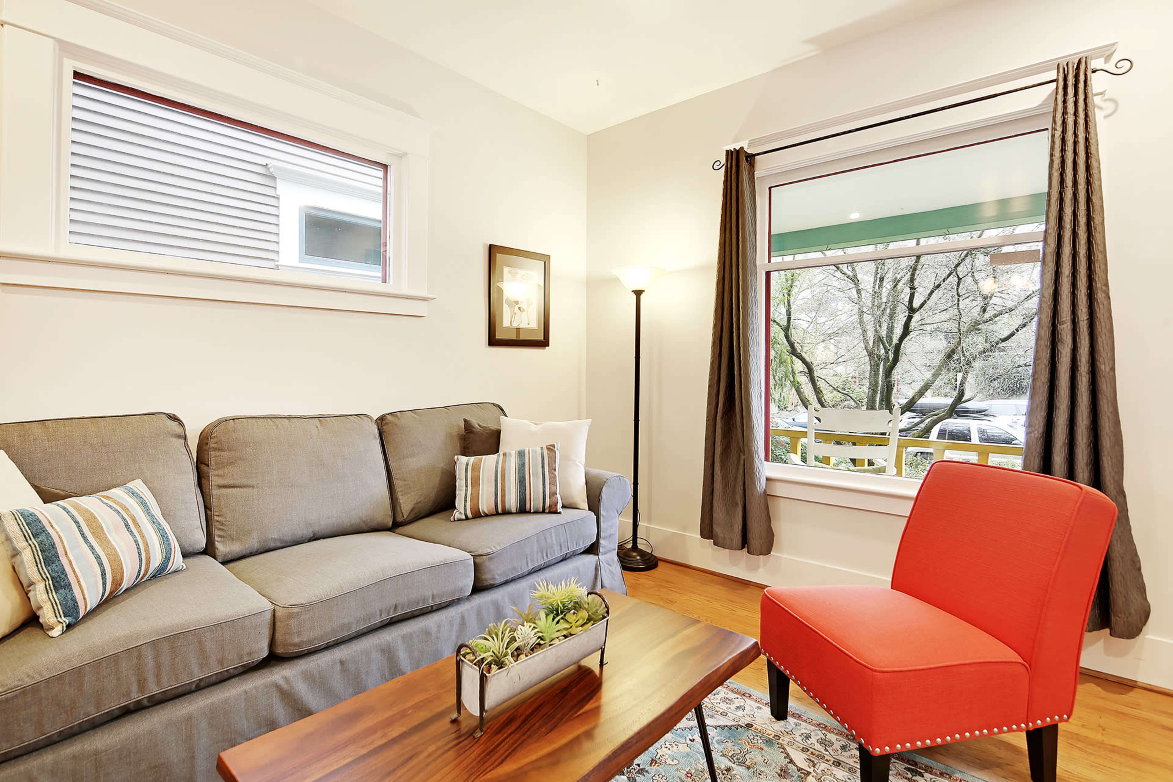 A gray couch with decorative pillows and a red chair sits next to a wooden coffee table in a well-lit living room with large windows.
