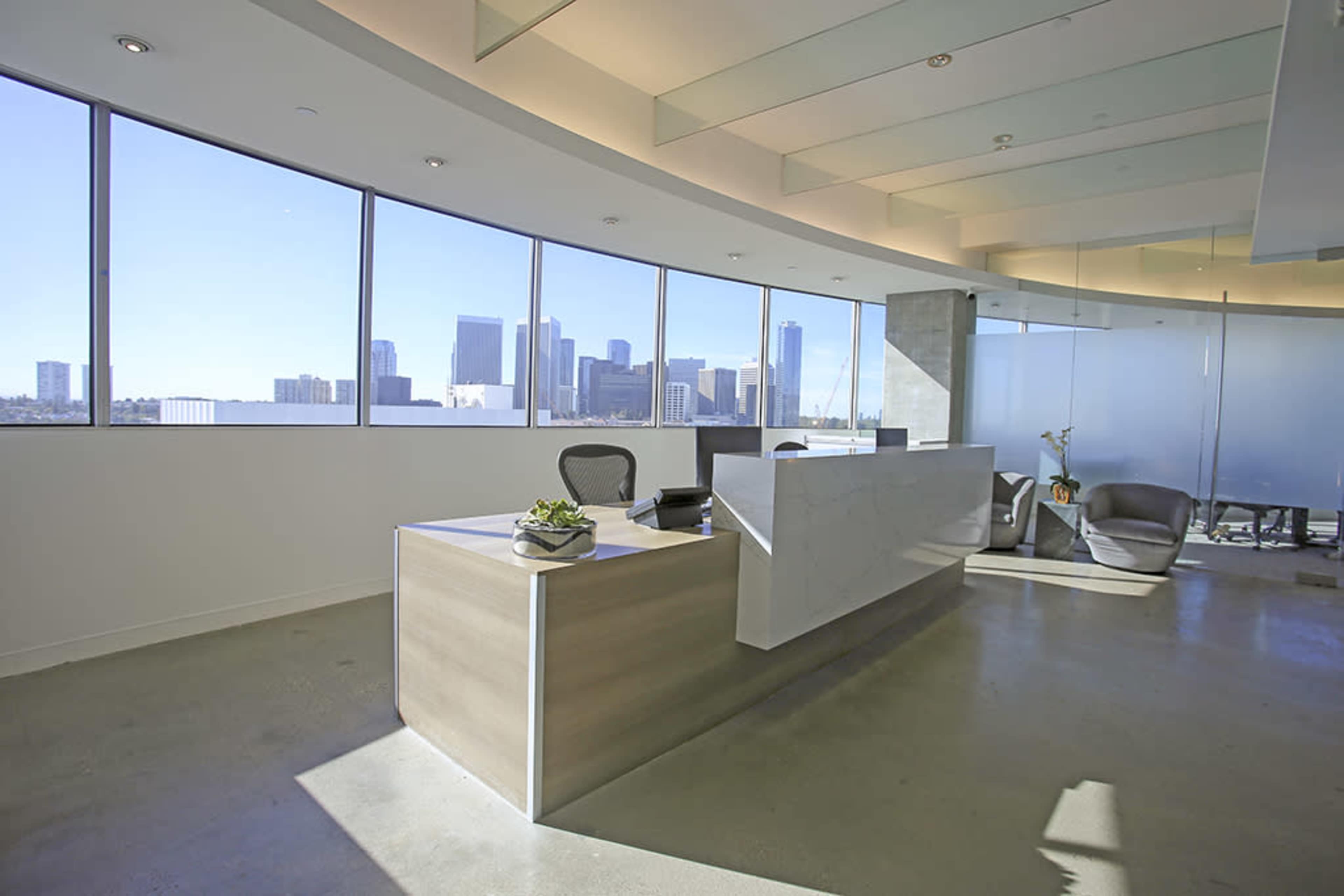 A modern office reception area features large windows overlooking a city skyline, with a light-colored desk and seating arrangement.