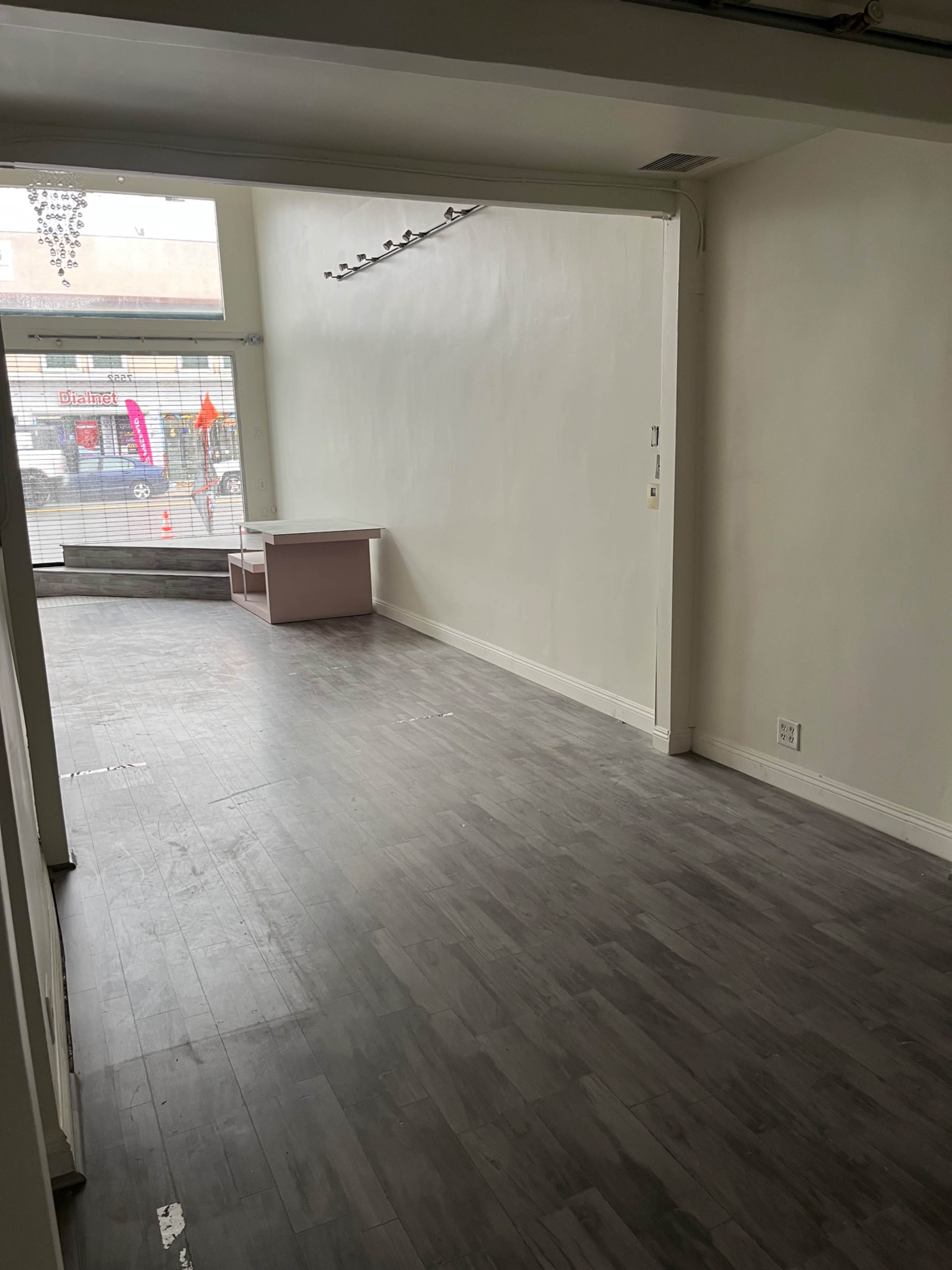 The image shows an empty retail space with light-colored walls, a wooden floor, and a small counter near the front window.