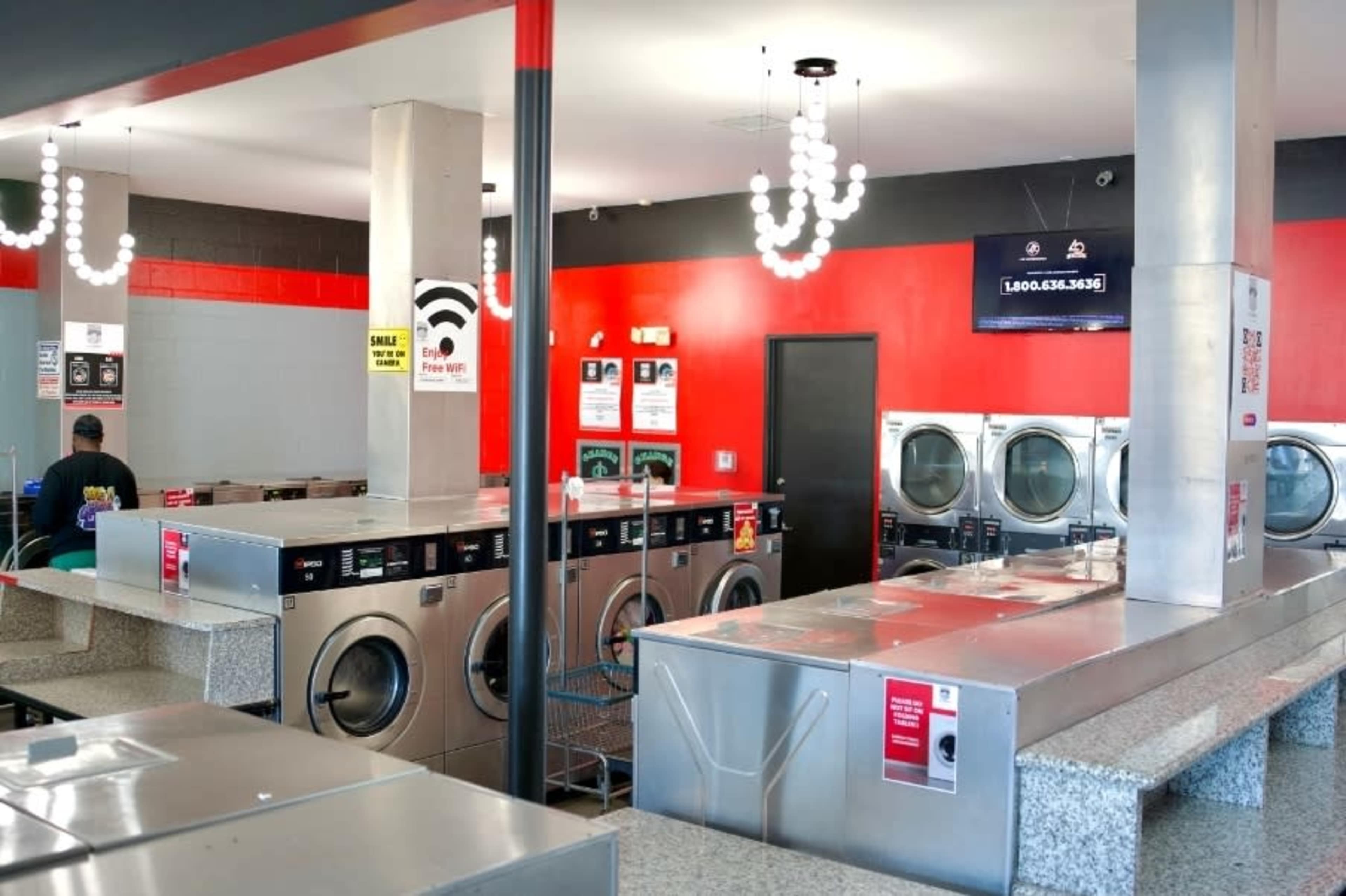 The image shows a modern laundromat with stainless steel machines, a granite folding area, and bright red accent walls.