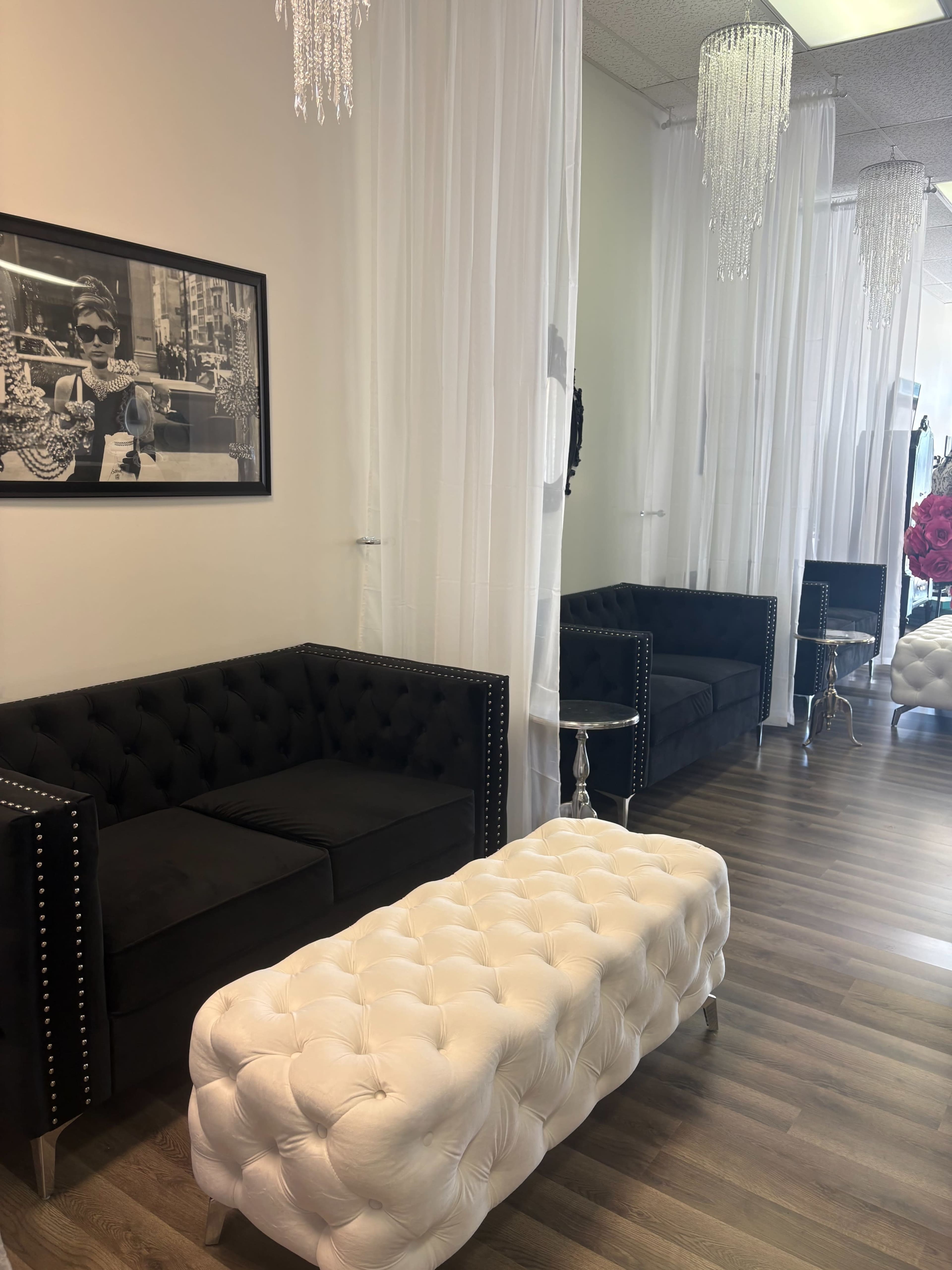 The image shows a modern waiting area featuring two black upholstered couches, a white tufted ottoman, and crystal pendant lights hanging from the ceiling.