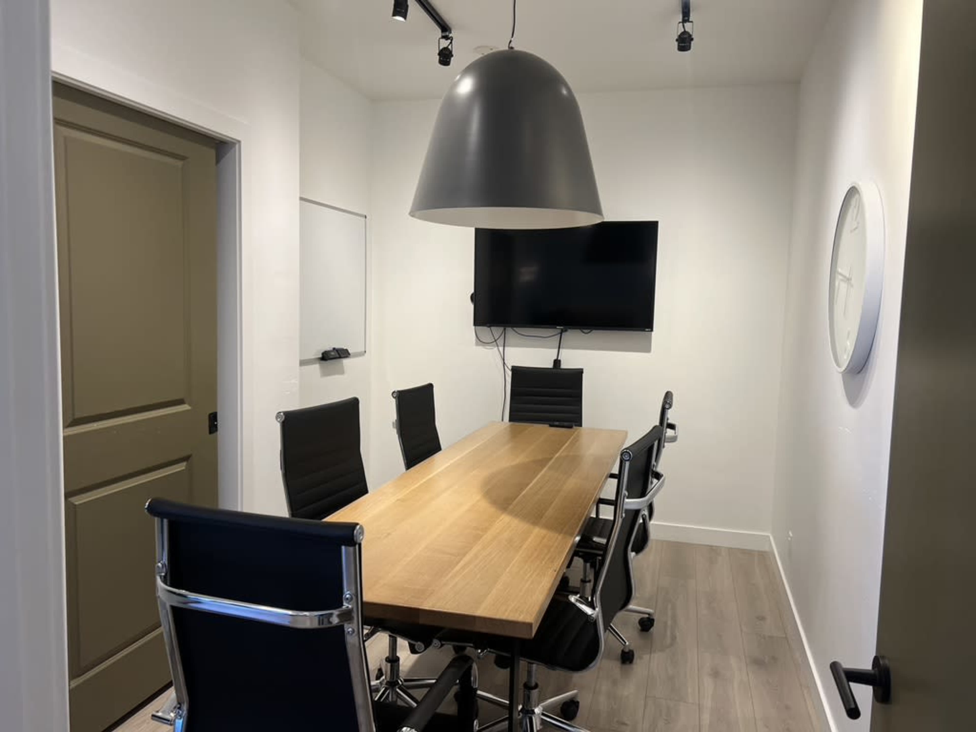 The image shows a meeting room featuring a long wooden table surrounded by six black chairs, with a large gray pendant light and a wall-mounted television.