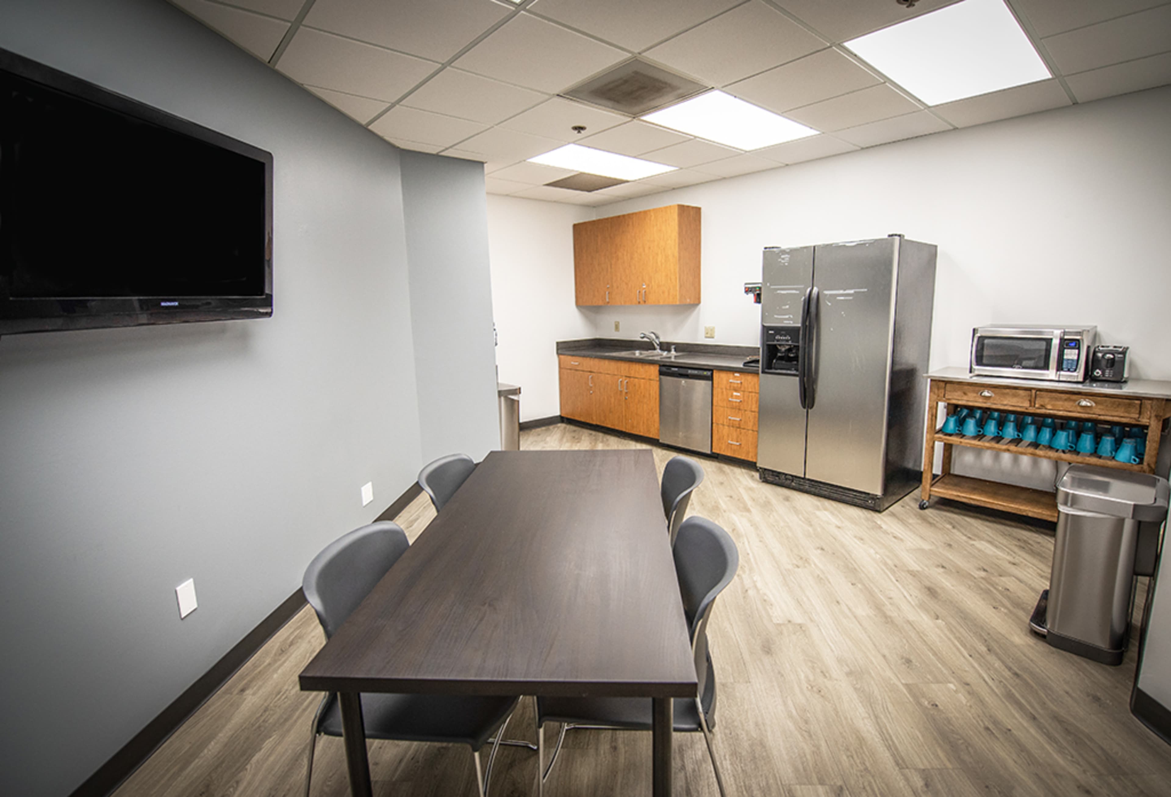 A kitchen area with a dining table, chairs, a refrigerator, a microwave, and wooden cabinets against a gray wall.