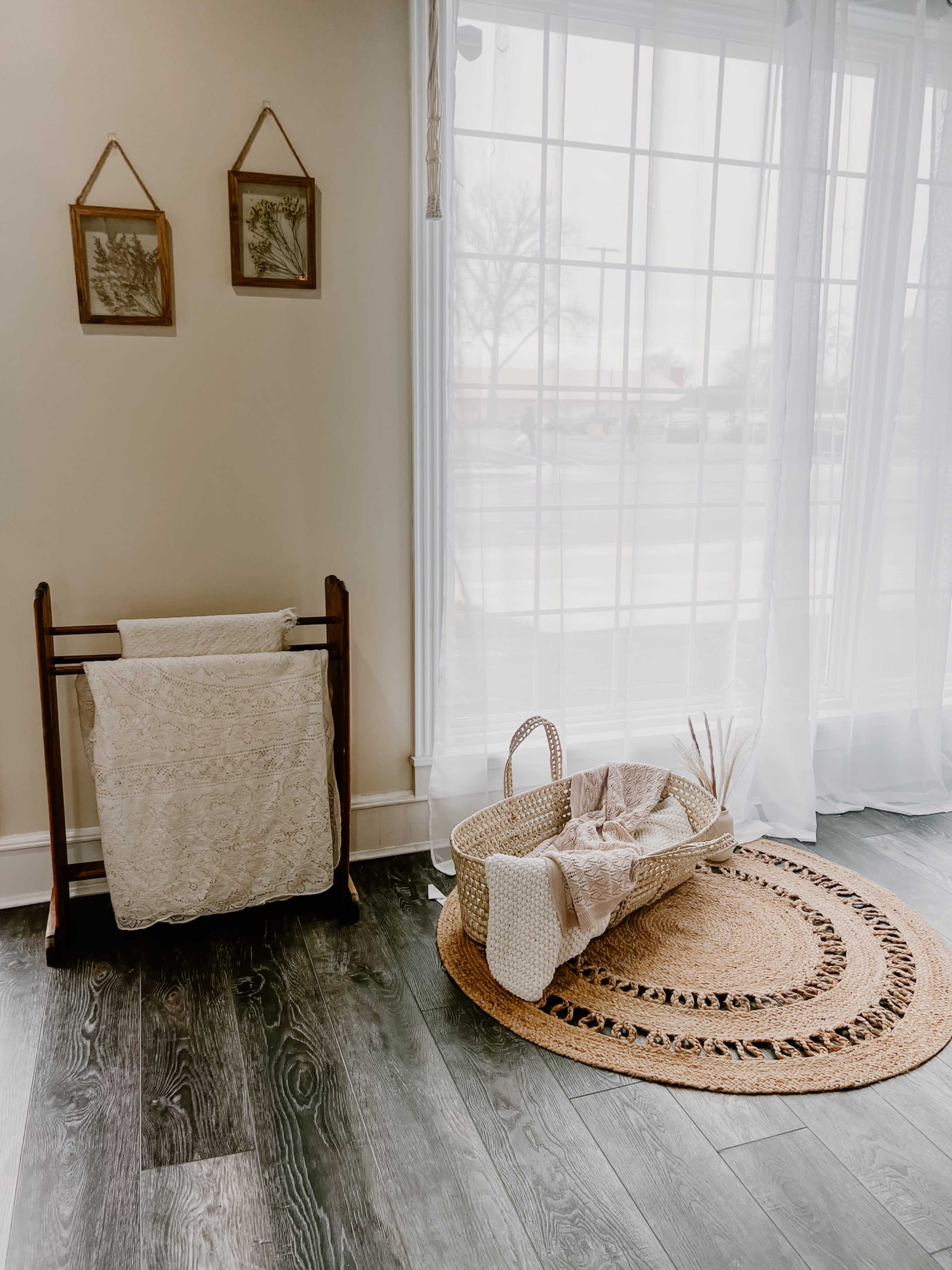 A wicker basket is placed on a round jute rug next to a wooden linen rack and sheer curtains letting in natural light.