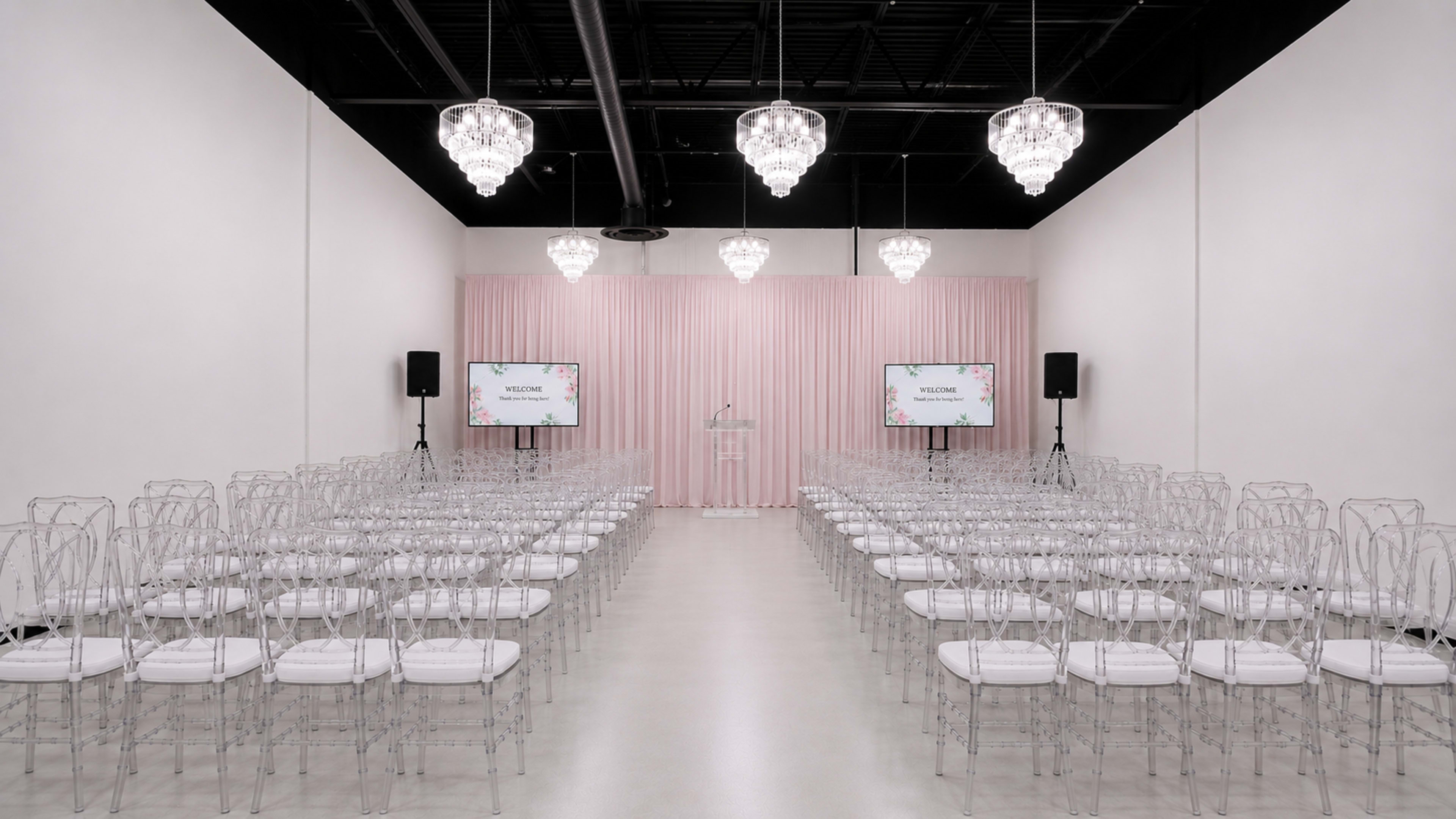 The image shows a modern event space set up for a ceremony, featuring rows of transparent chairs facing a clear podium with screens on either side.