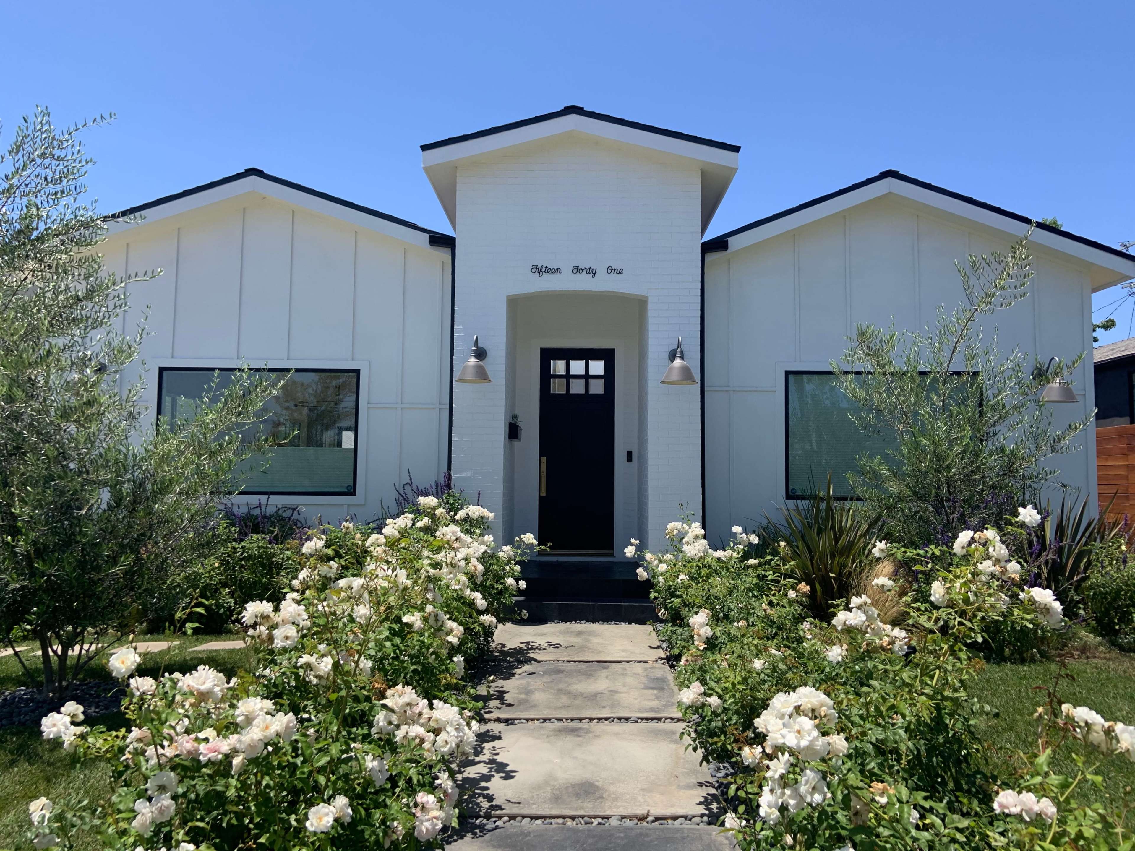 A modern white house with a geometric facade, flanked by flowering shrubs and plants, features a black front door and large windows.