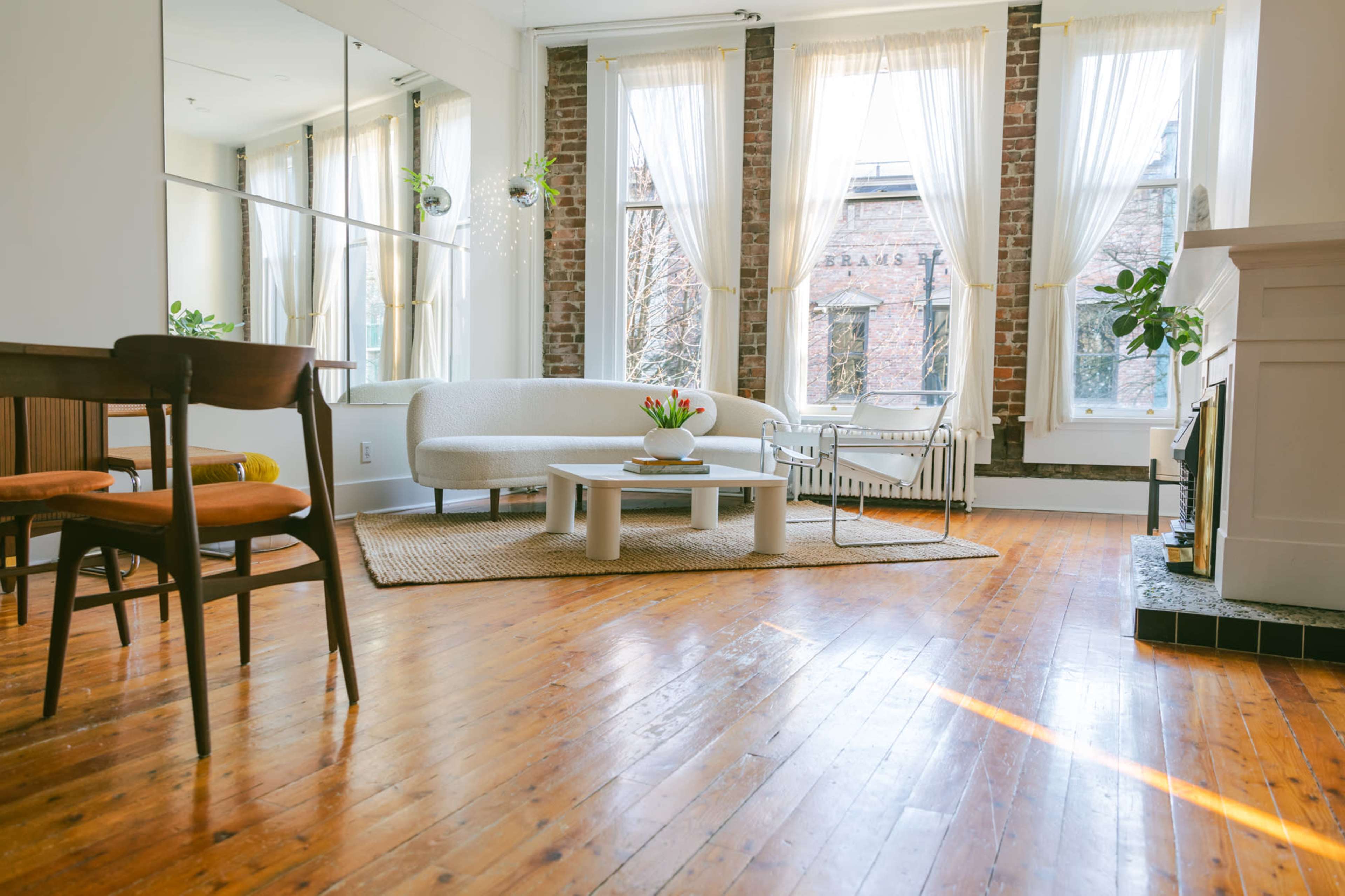 A bright living room features a curved white sofa, a round coffee table, and large windows with sheer curtains, all set on polished wooden floors.