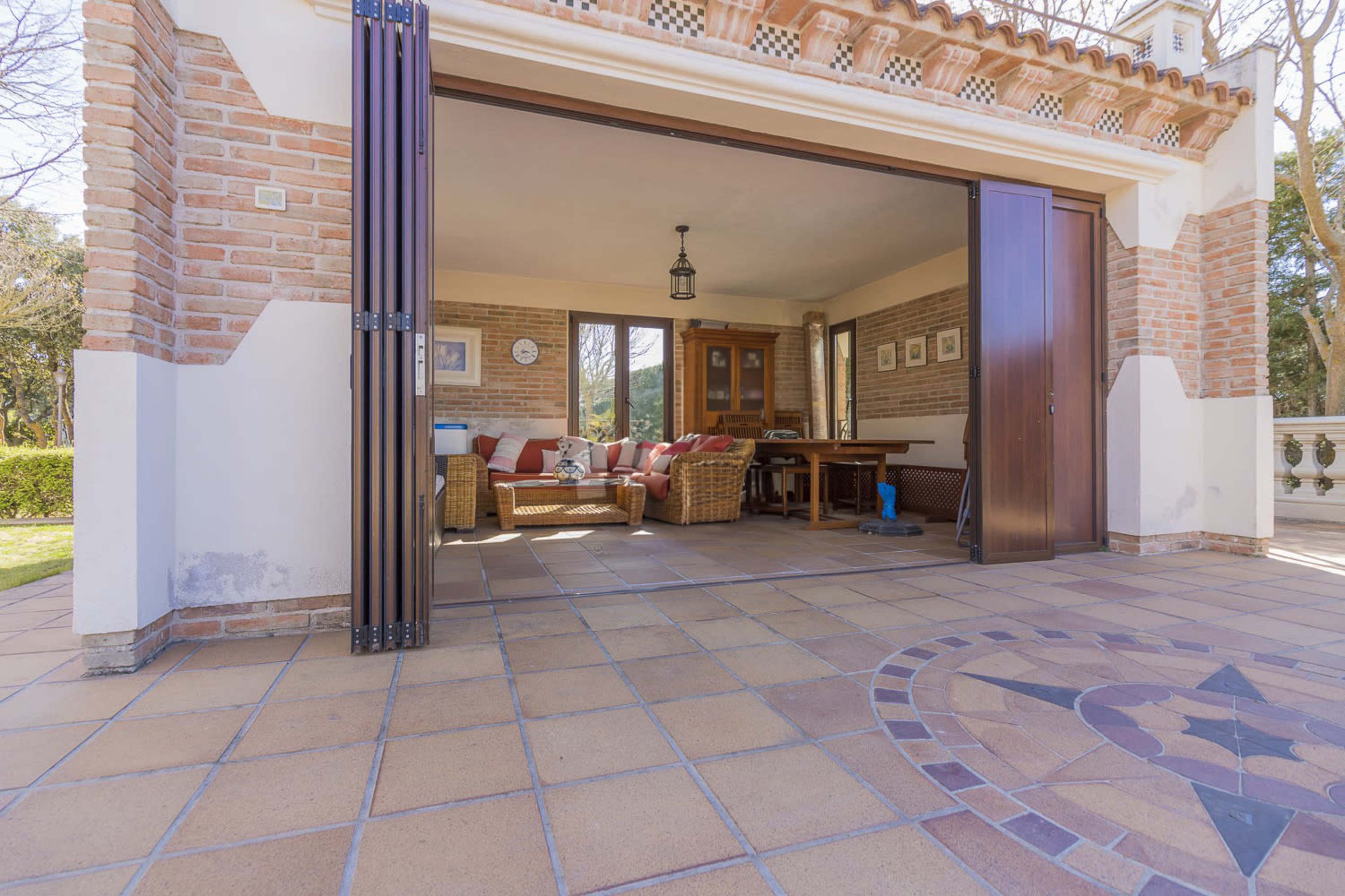 The image shows a spacious open indoor area with large sliding doors leading to an outdoor patio, featuring a table and seating arrangement.