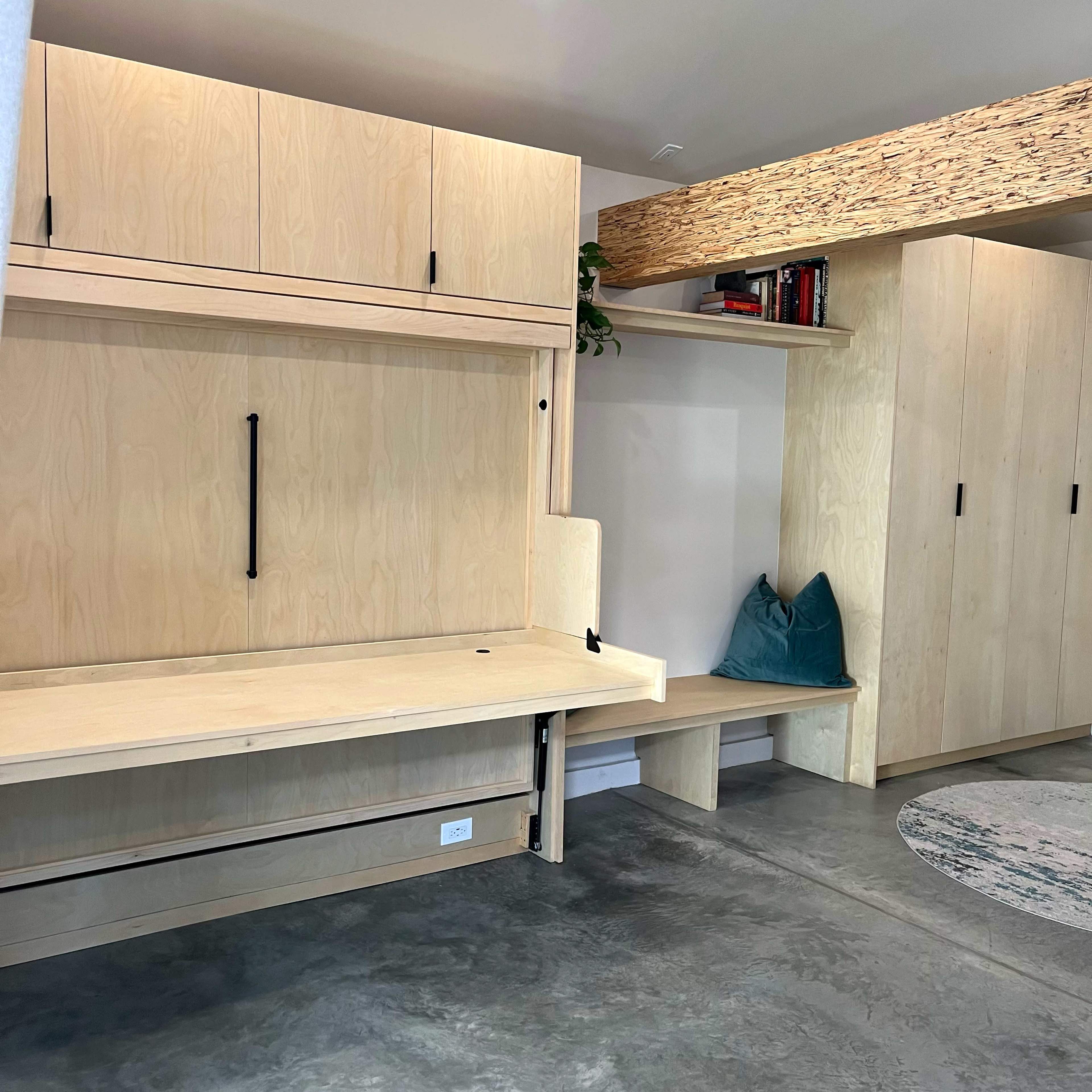 The image shows a minimalist interior featuring a light wood wall-mounted desk that folds down, with adjacent built-in cabinets and a small seating area.