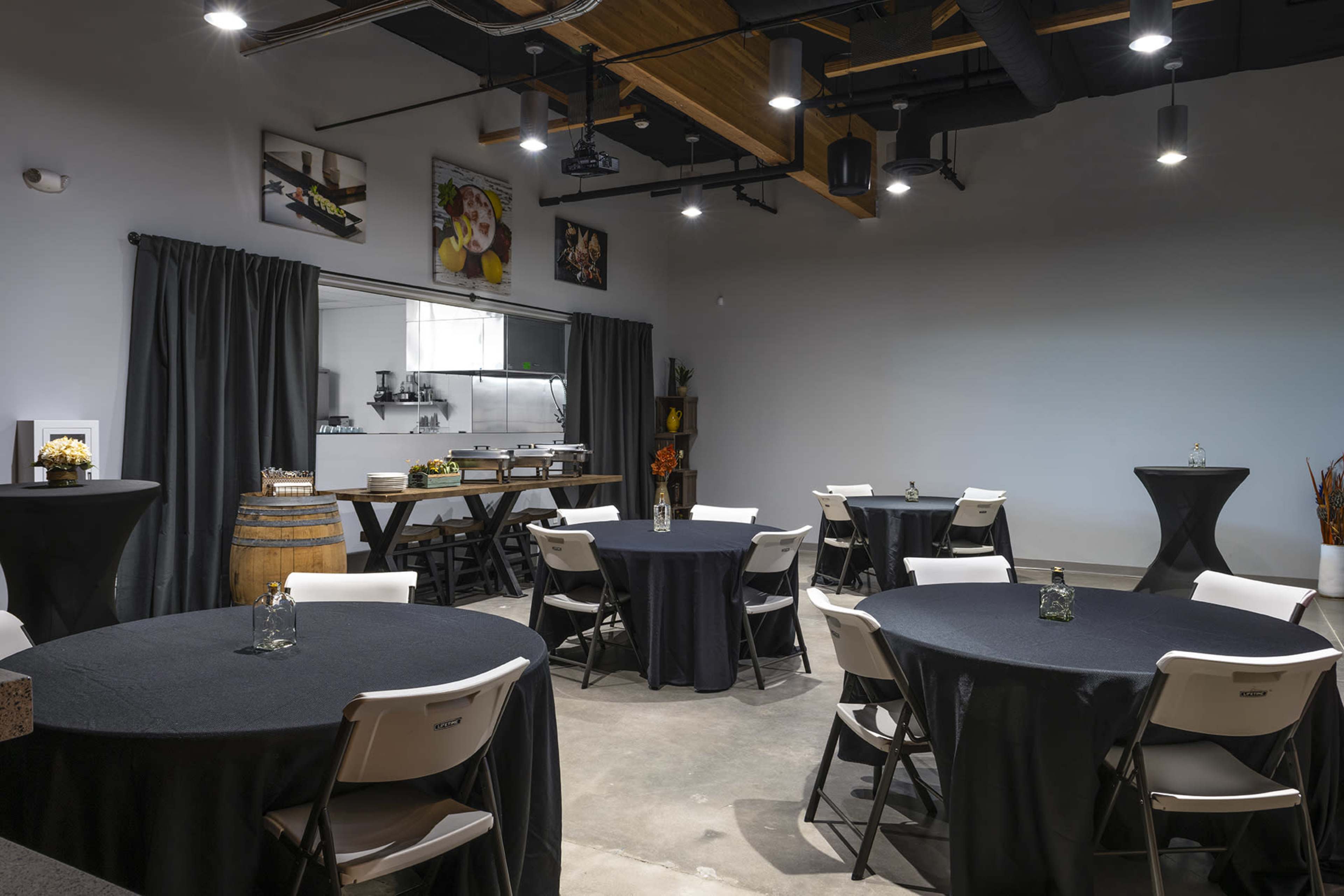 A spacious event room features round tables covered with black tablecloths, a high table, and a large barrel, with a kitchen area visible in the background.