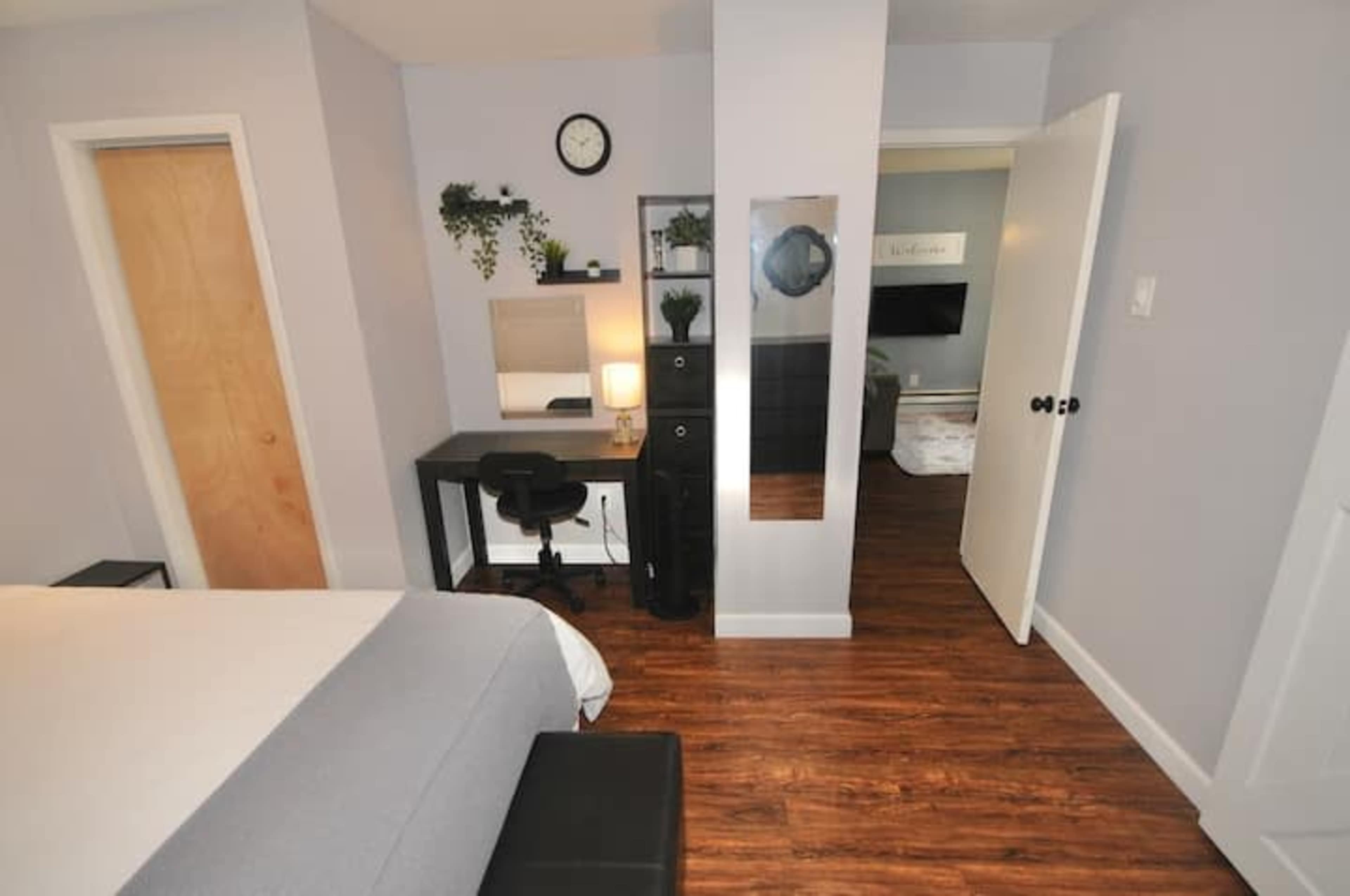 A small bedroom featuring a bed, a desk with a chair, a mirror, and a TV visible through an open door.