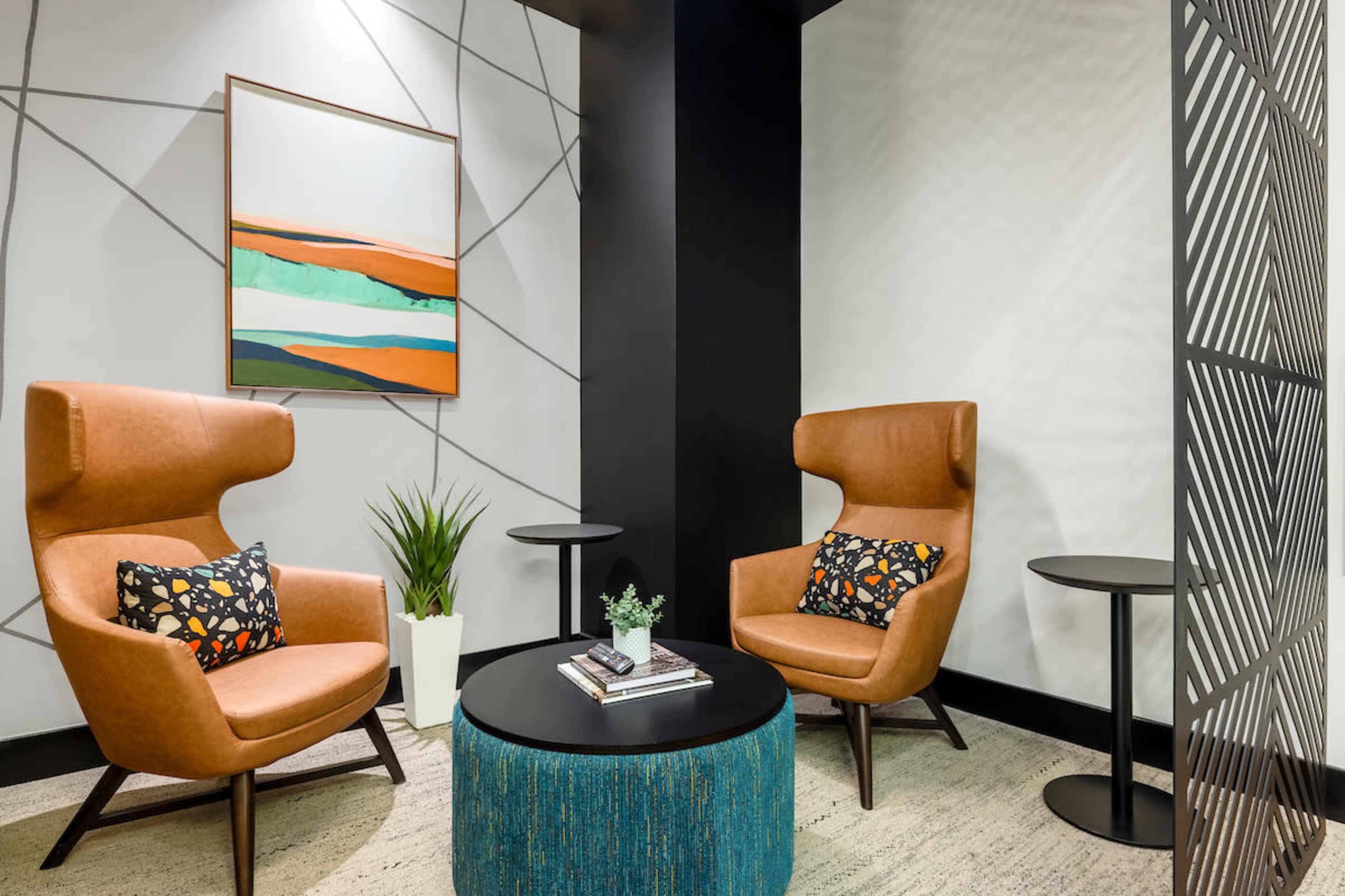 A modern seating area featuring two brown armchairs with decorative pillows, a round black coffee table, and a small green plant, set against a backdrop of geometric wall patterns and artwork.