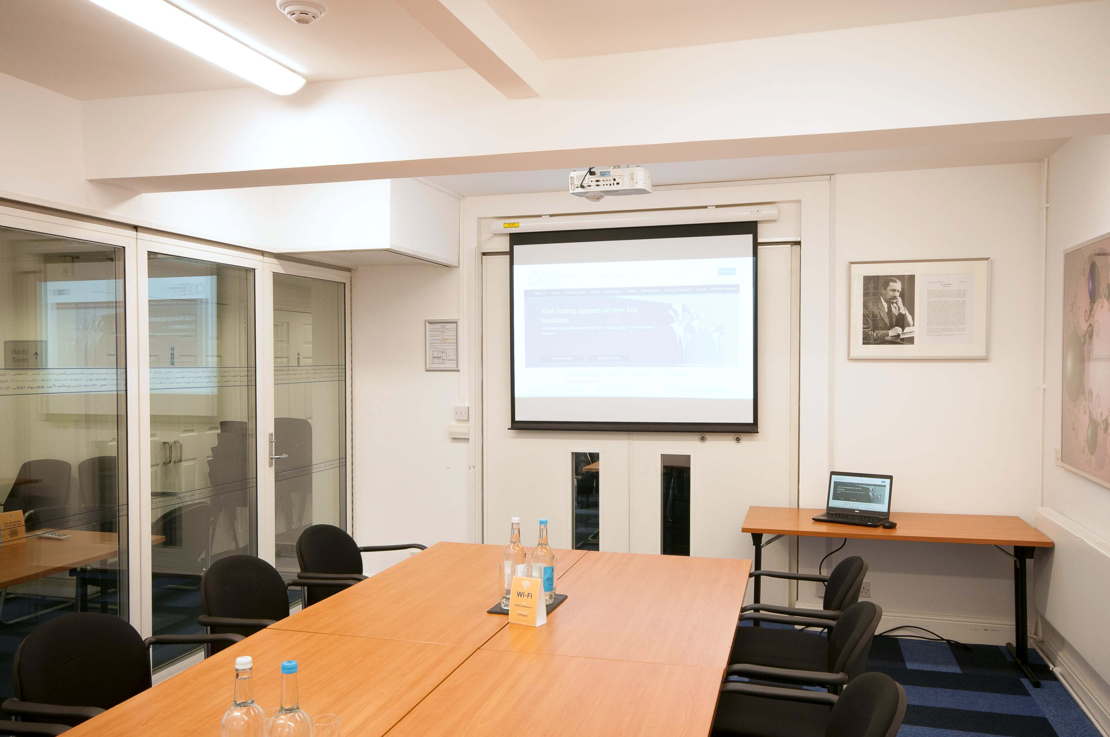 A conference room is set up with a large wooden table, black chairs, a projector screen displaying a website, and a laptop on the side.
