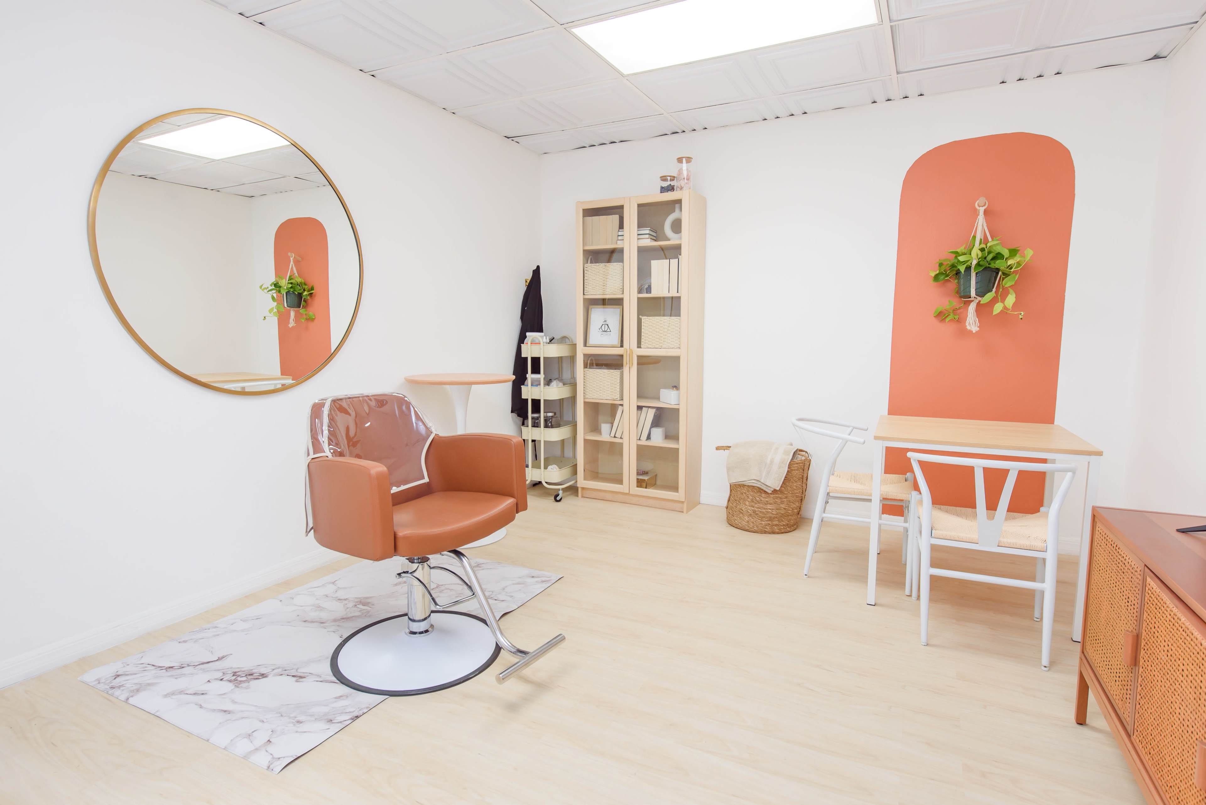 A modern room featuring a circular mirror, a hair styling chair, a wooden table with chairs, a shelving unit, and decorative plants against a light-colored wall.