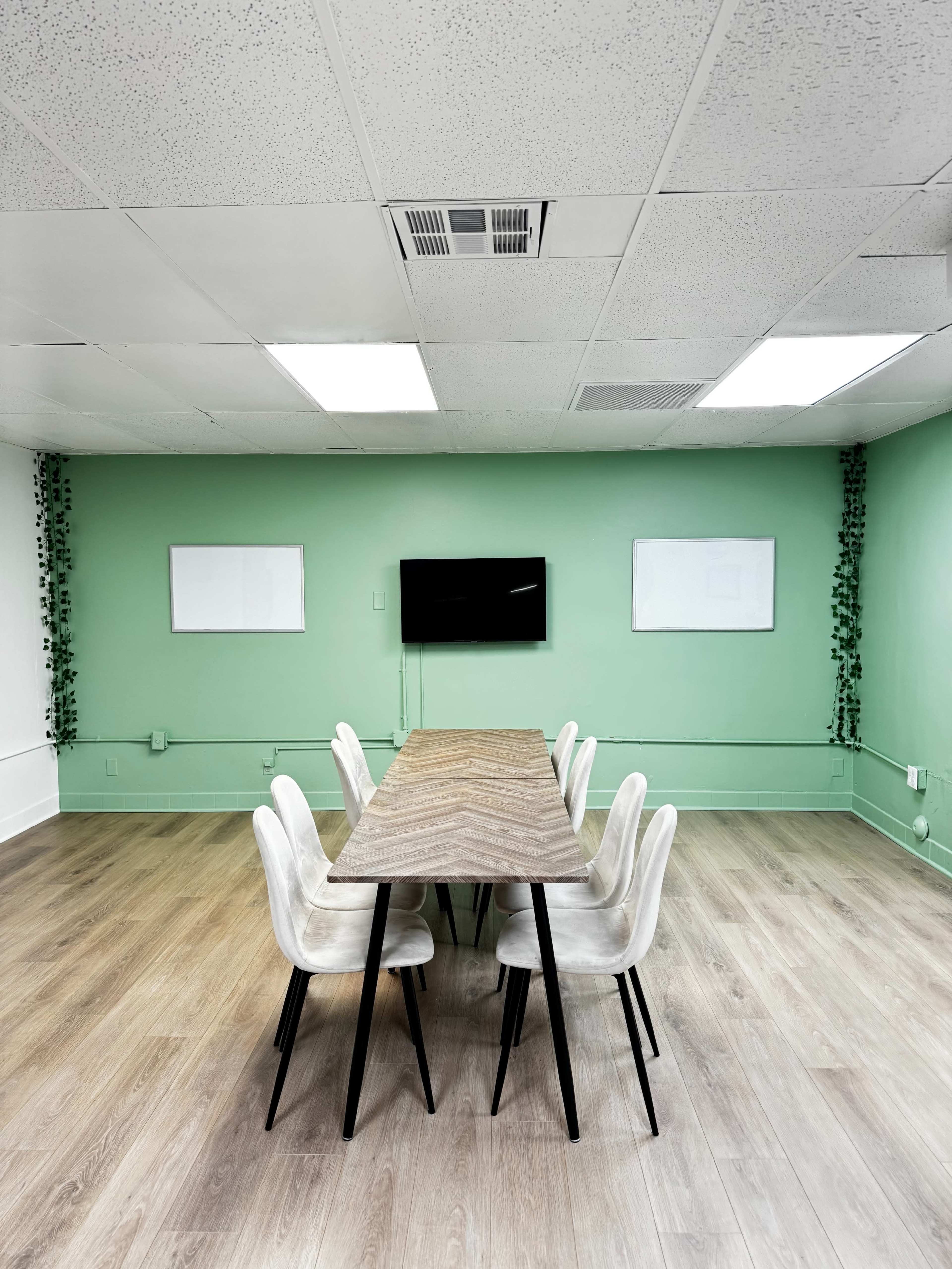 The image shows a conference room with a long wooden table surrounded by white chairs, green walls, a television mounted on the wall, and two blank whiteboards.