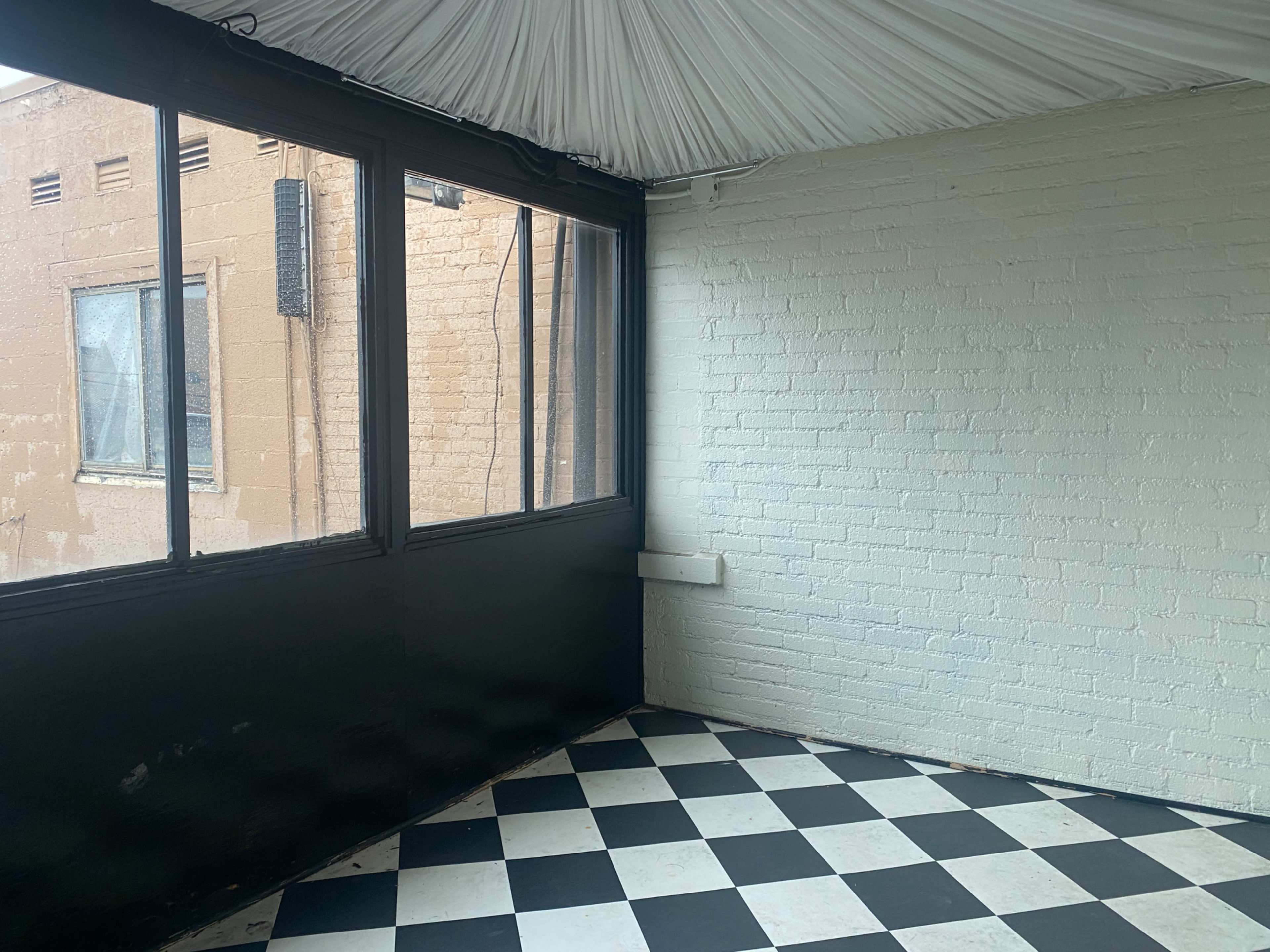An empty room featuring a black and white checkered floor, large windows with a view of a brick wall, and a ceiling covered with fabric.