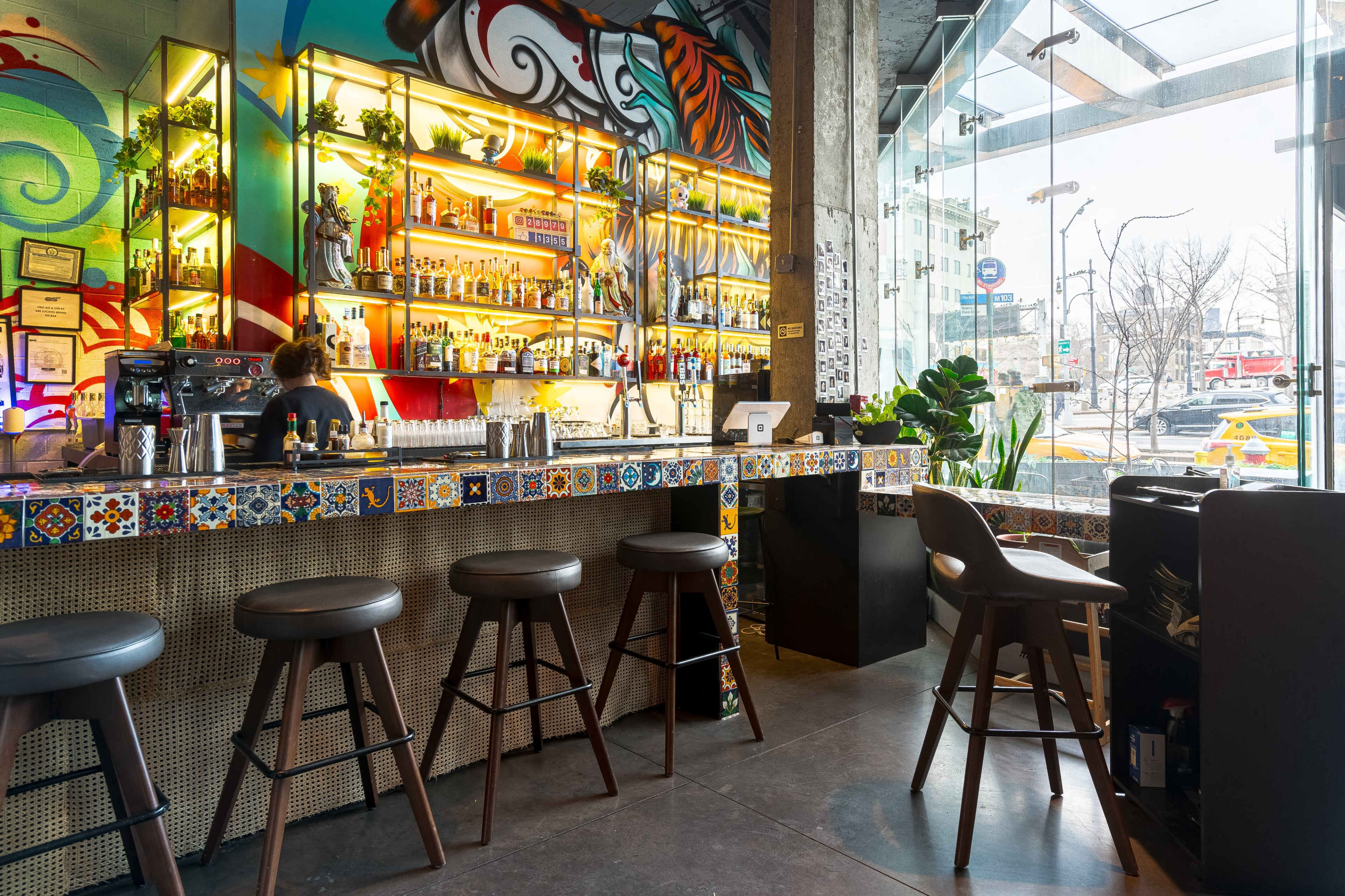 A vibrant bar with a colorful mural, lined with shelves of drinks and several bar stools arranged in front.
