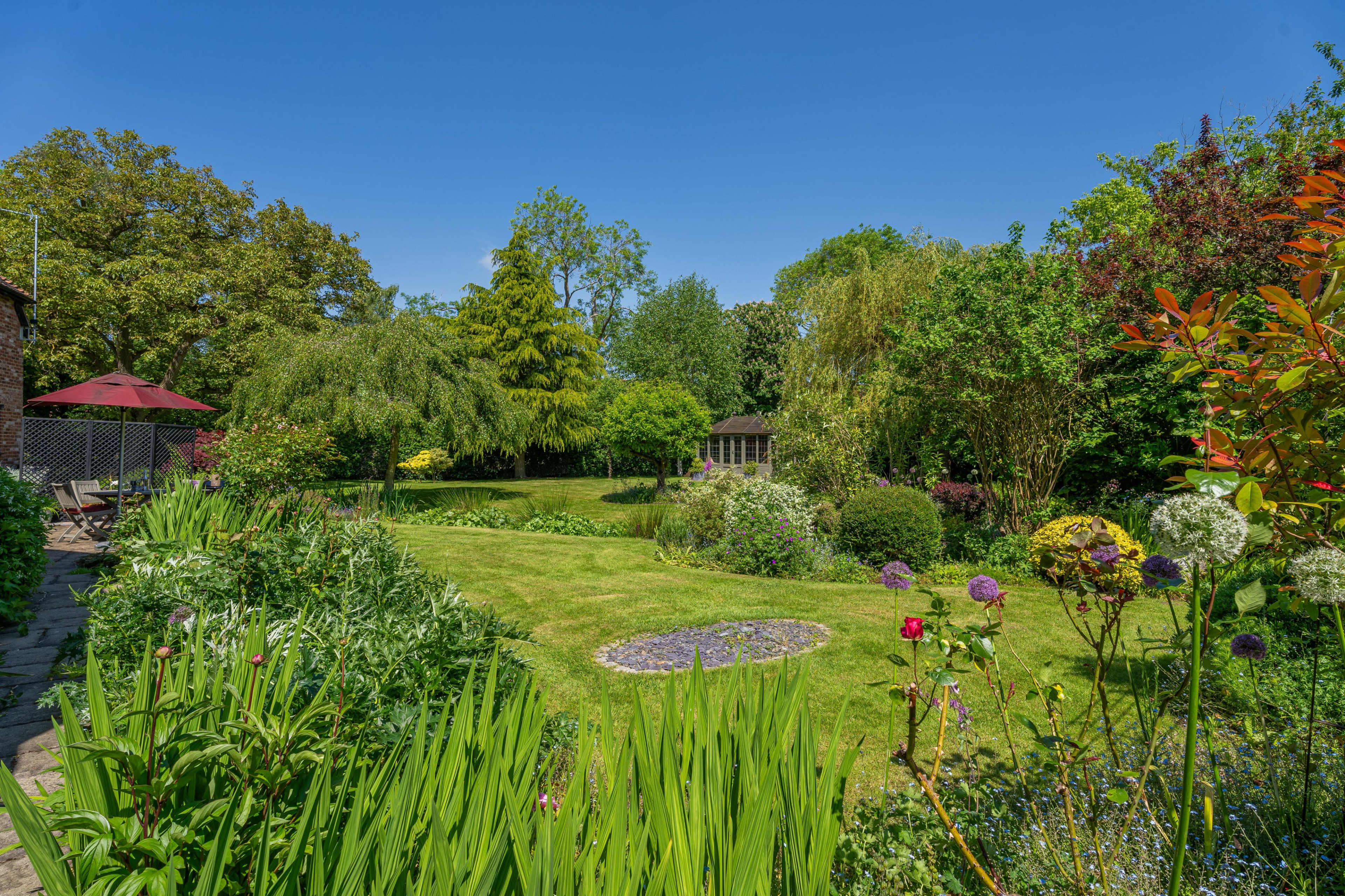 A well-maintained garden features a variety of plants and trees, with a circular stone path and a wooden shed in the background.
