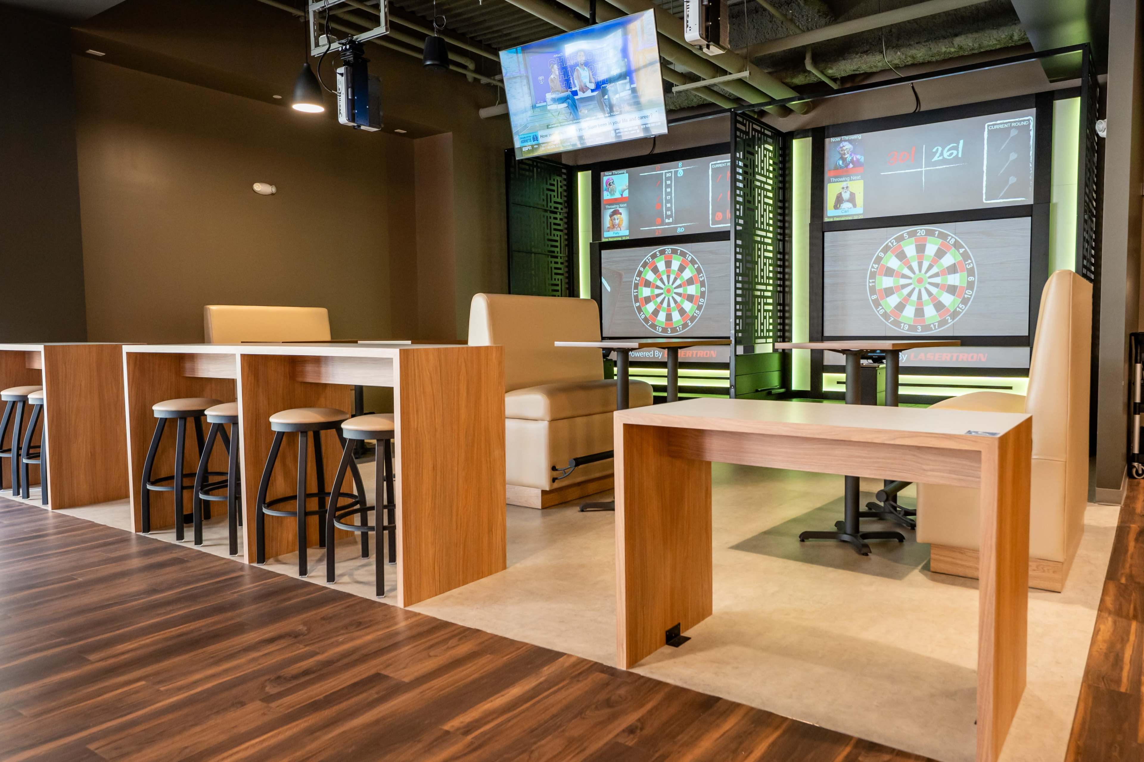 The image shows a modern recreational space featuring dartboards projected on screens, sleek seating arrangements, and a bar area with high stools.