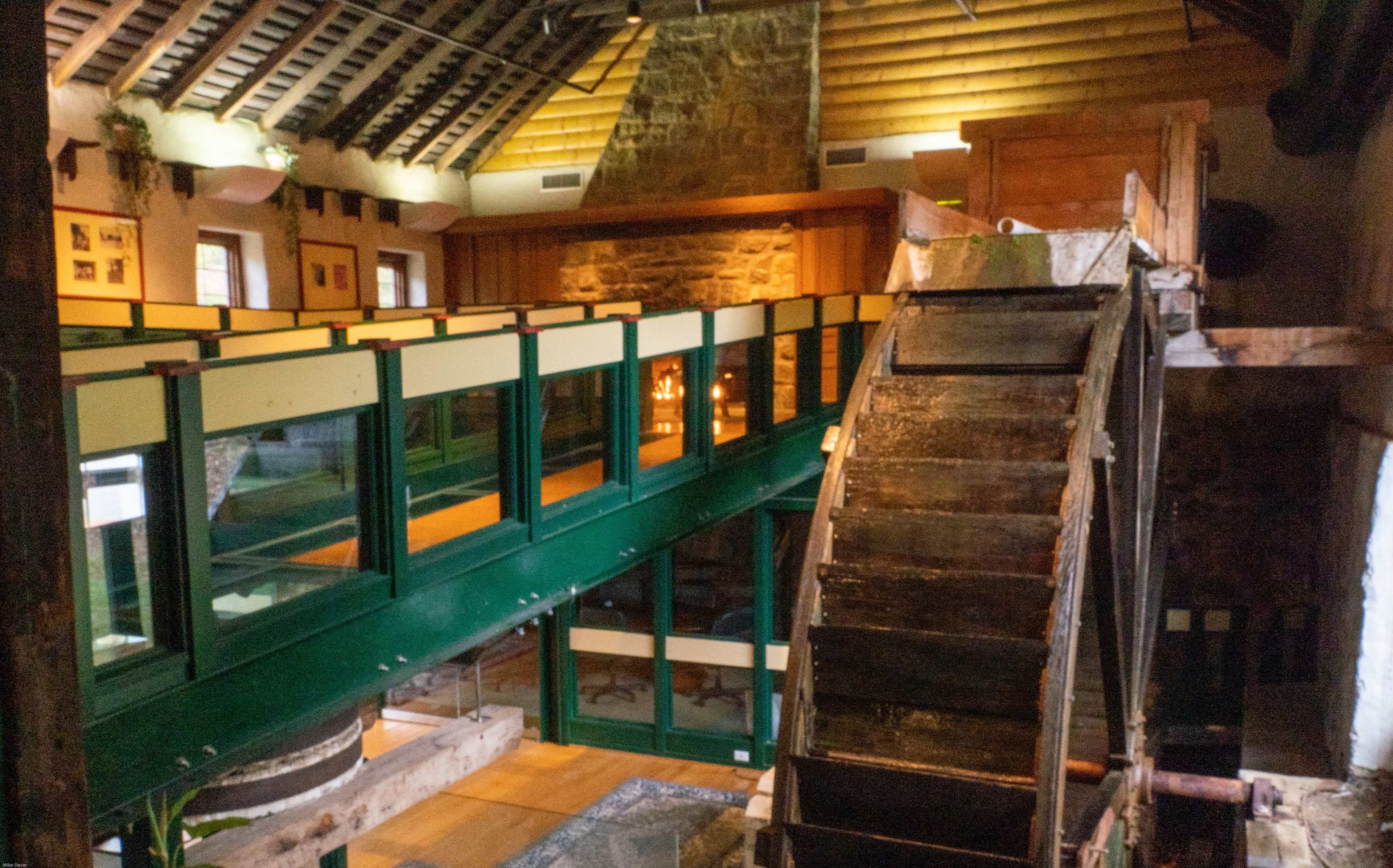 A rustic interior of a building with exposed wooden beams, featuring a large water wheel and a balcony overlooking the main space.