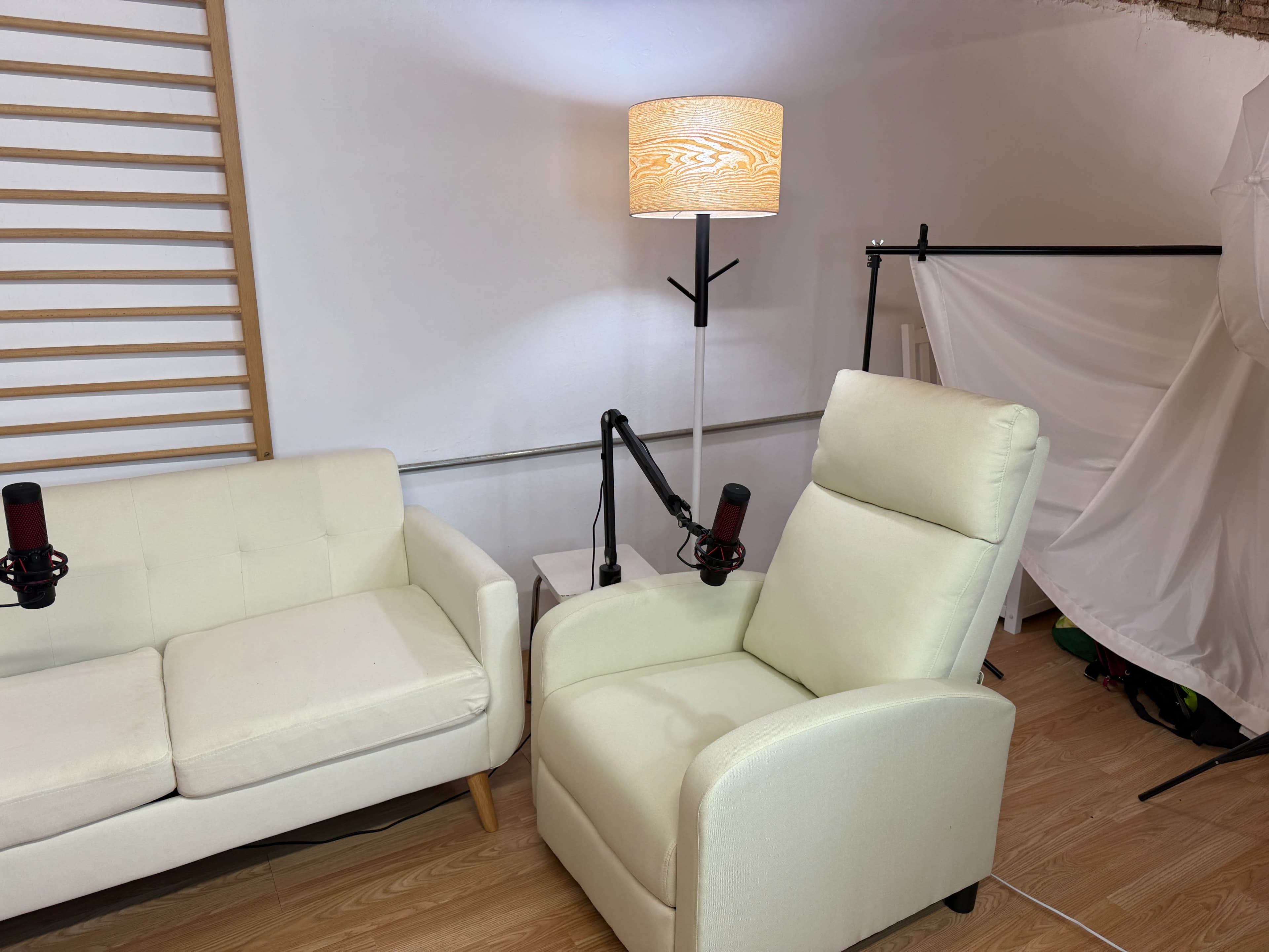 The image shows a cozy indoor setting with a white couch, a reclining armchair, and a lamp, accompanied by microphones mounted on stands.