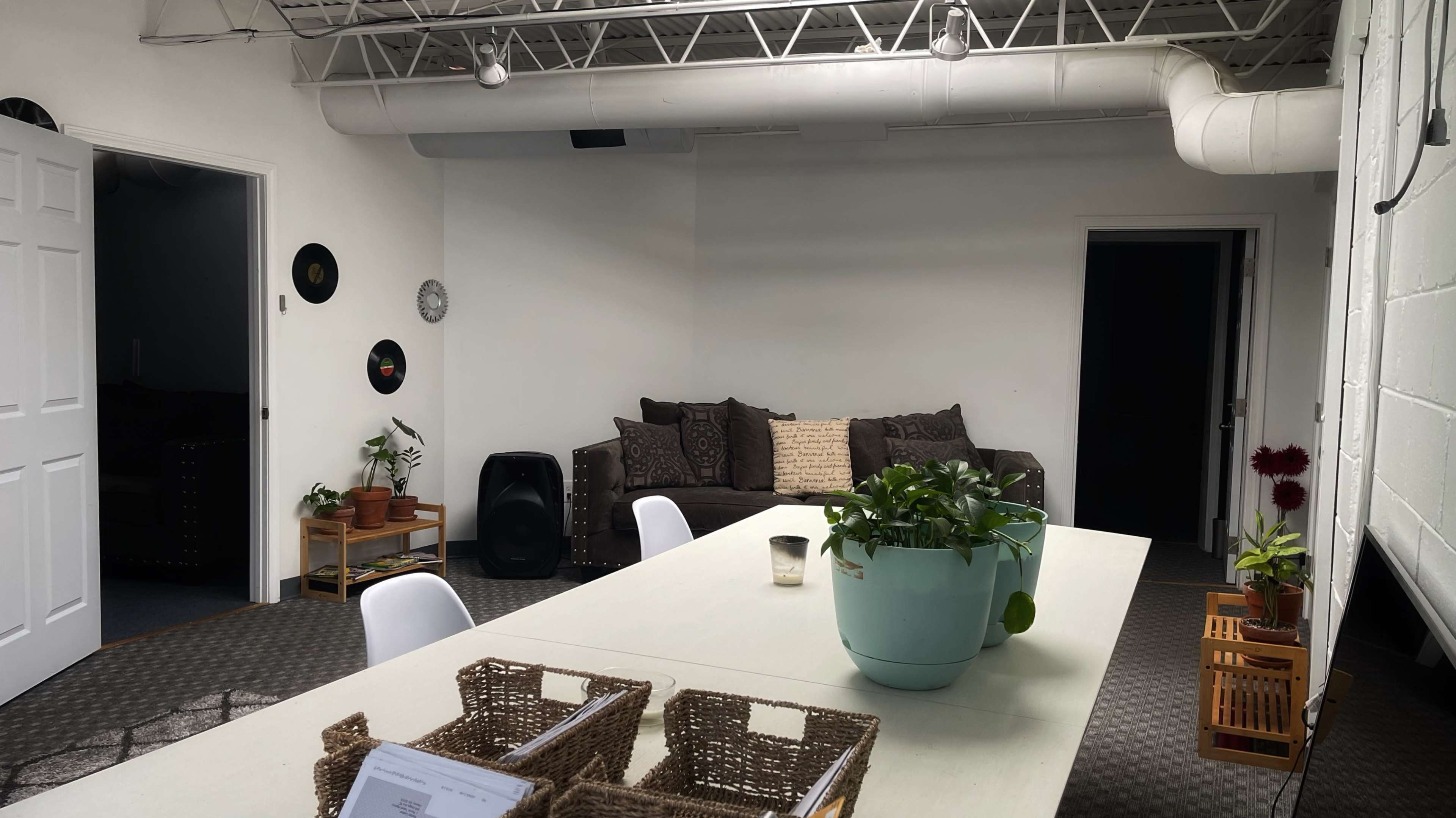 A modern, open room features a large table with plants, a couch along the wall, and a speaker in the corner.