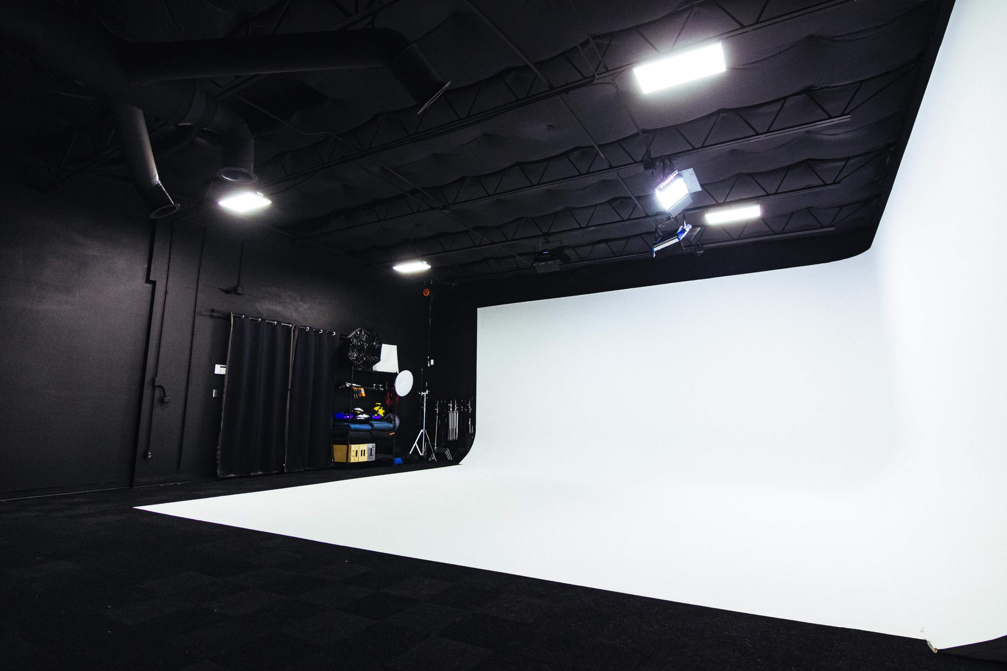 A photography studio features a large white seamless backdrop, bright overhead lights, and various equipment set against dark walls.