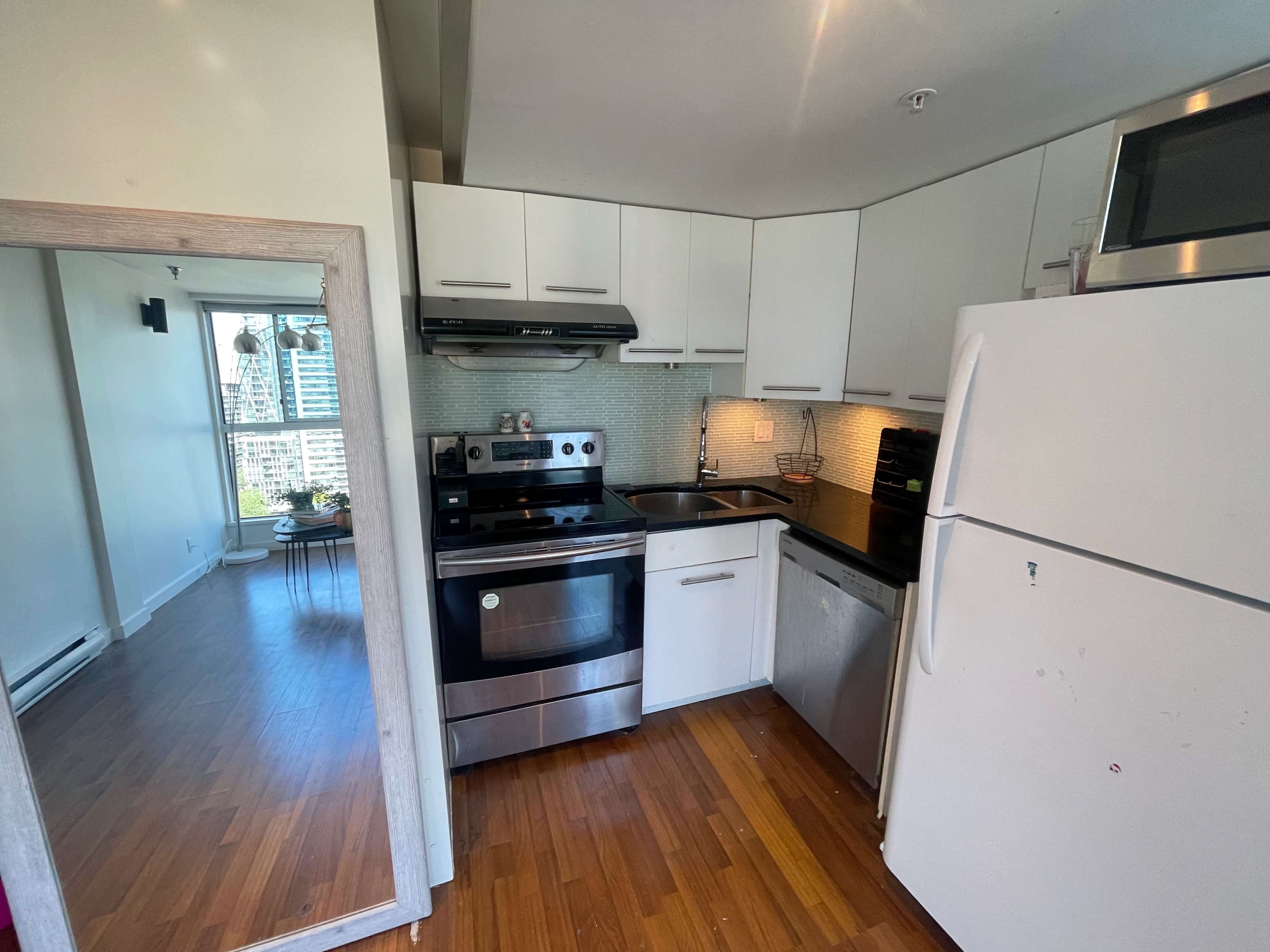 A modern kitchen with stainless steel appliances, white cabinets, and hardwood flooring, leading into a bright living space.