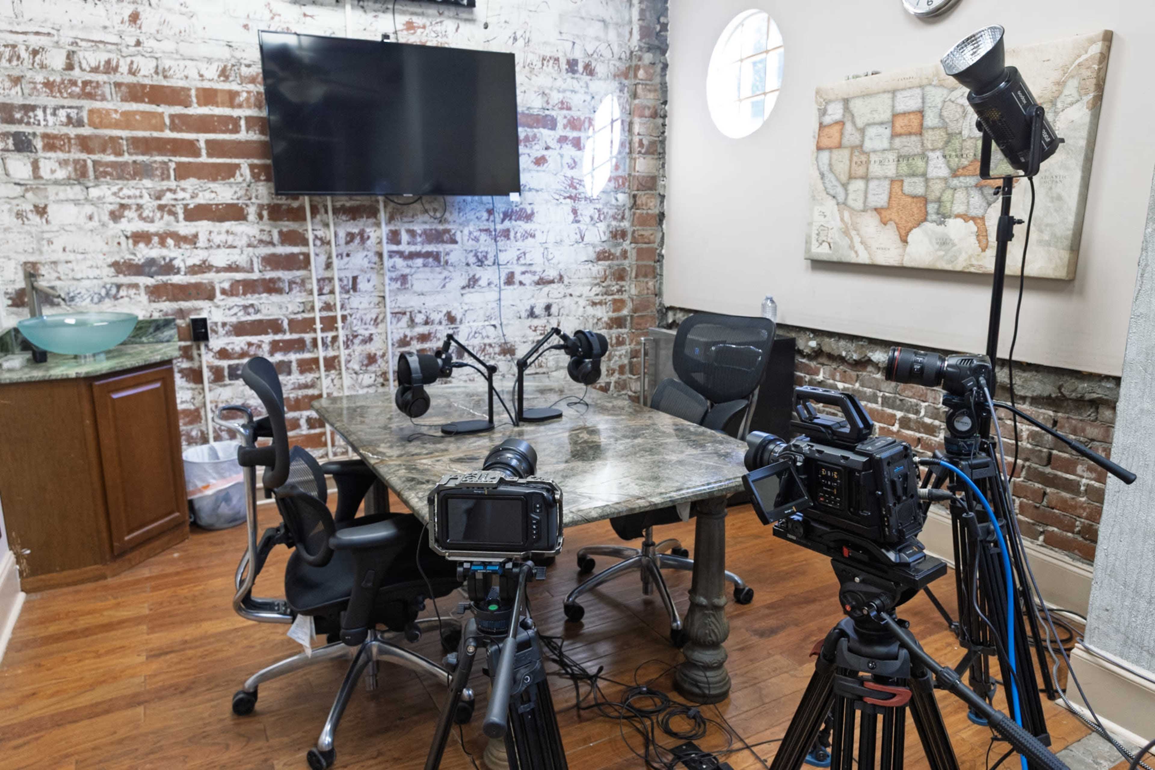 The image shows a brick-walled recording studio with a table and two microphones set up, surrounded by cameras and lighting equipment.