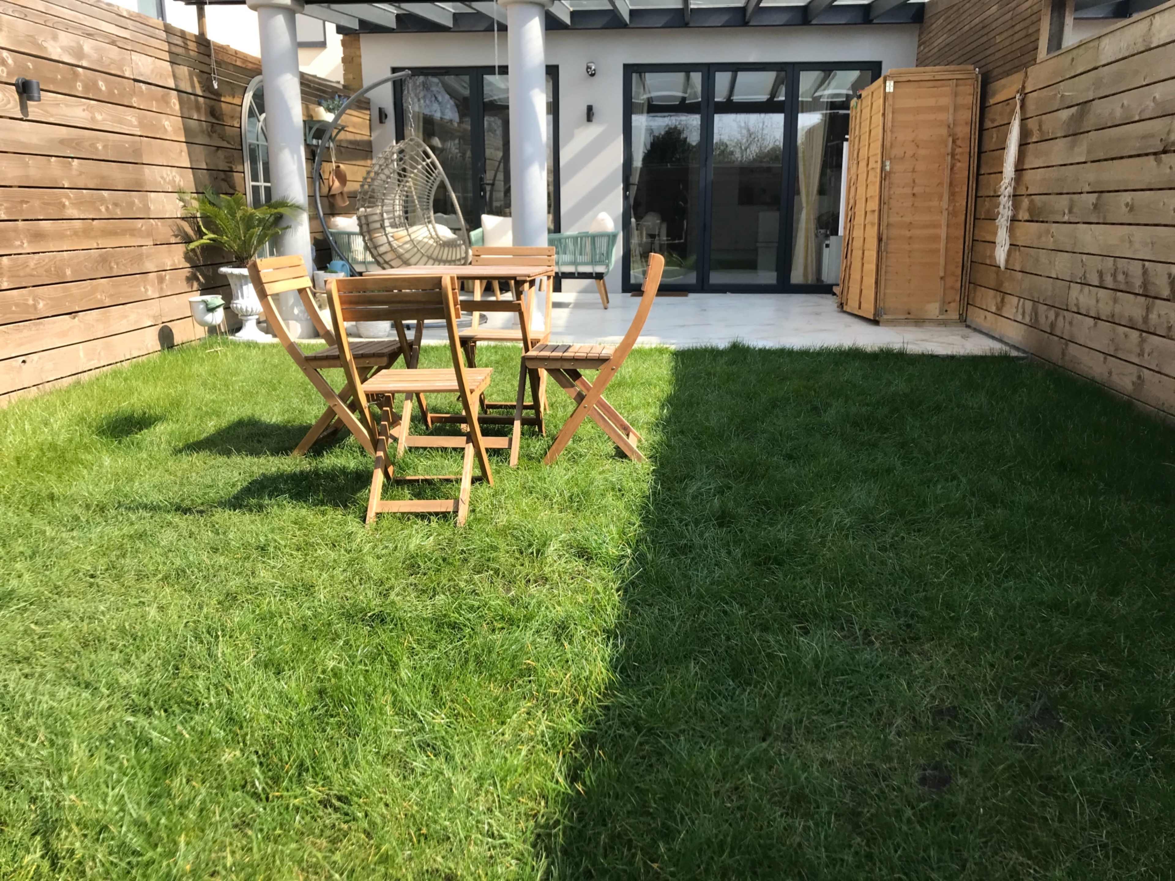 A wooden dining set with folding chairs is placed on a green lawn in a backyard, with a modern house in the background.