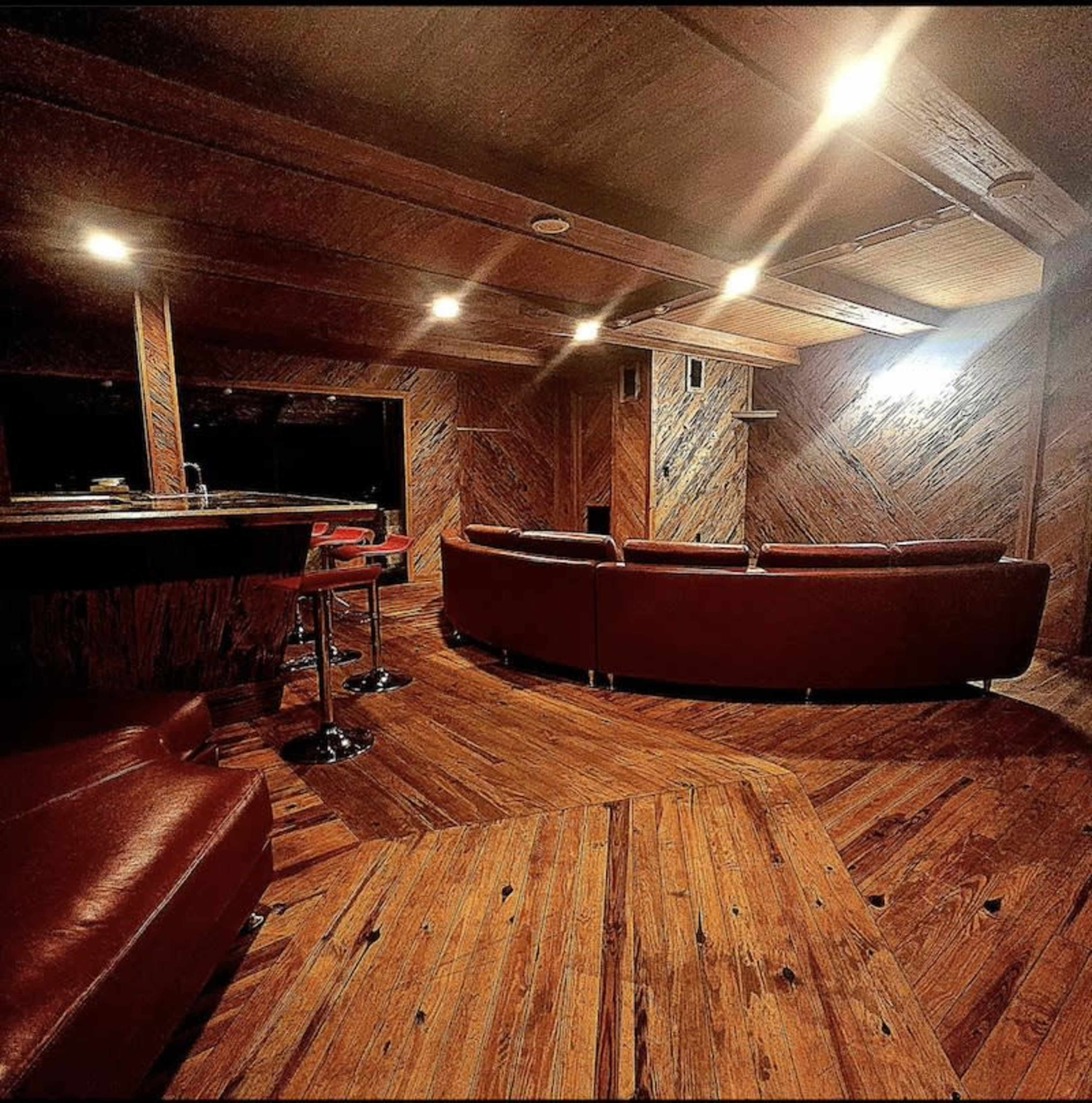 A spacious, dimly lit lounge with wooden flooring and walls, featuring a curved leather sofa and a bar area.