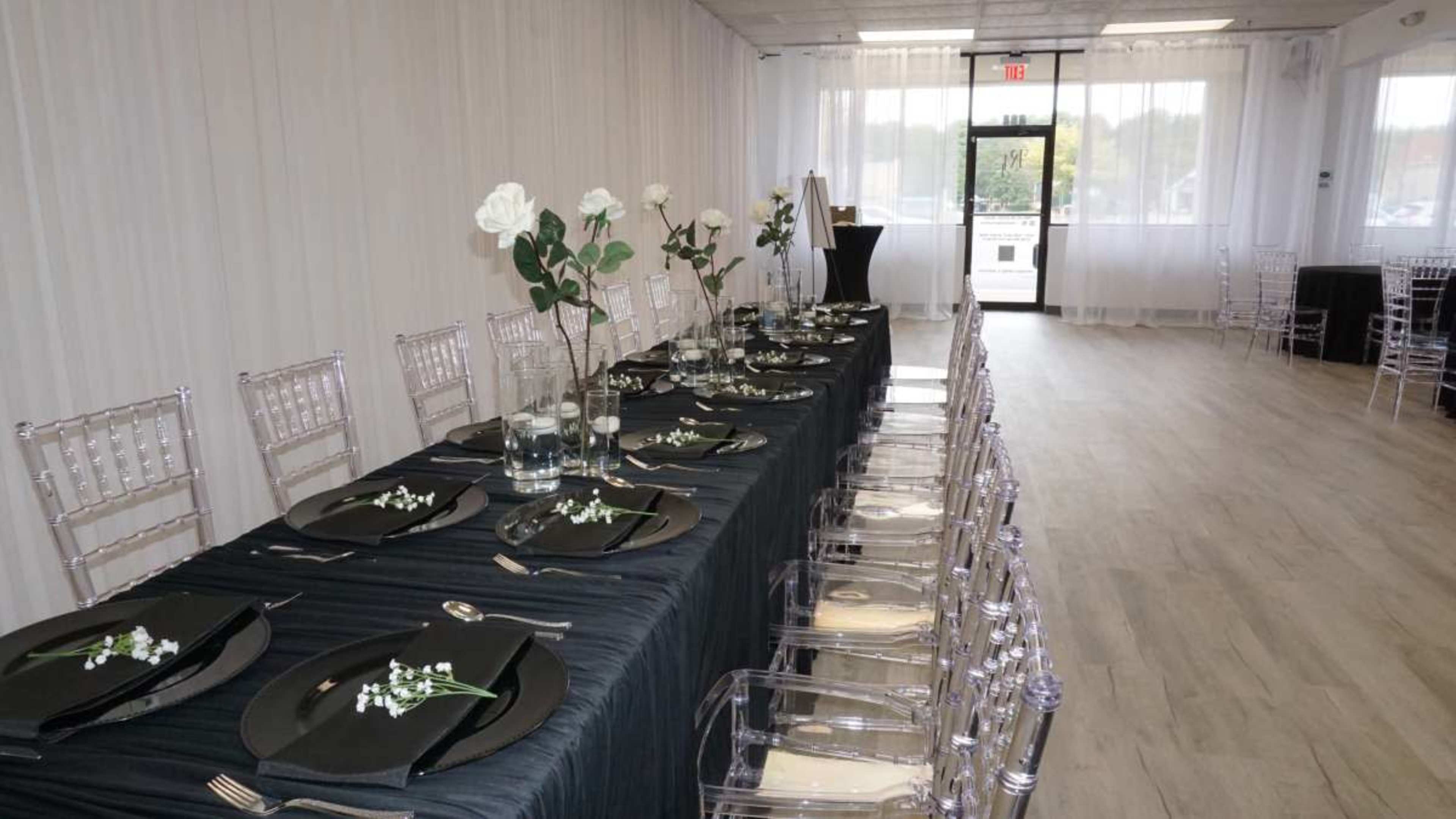 A long dining table is set with black tablecloths, clear ghost chairs, and decorative centerpieces of white roses in glass vases.