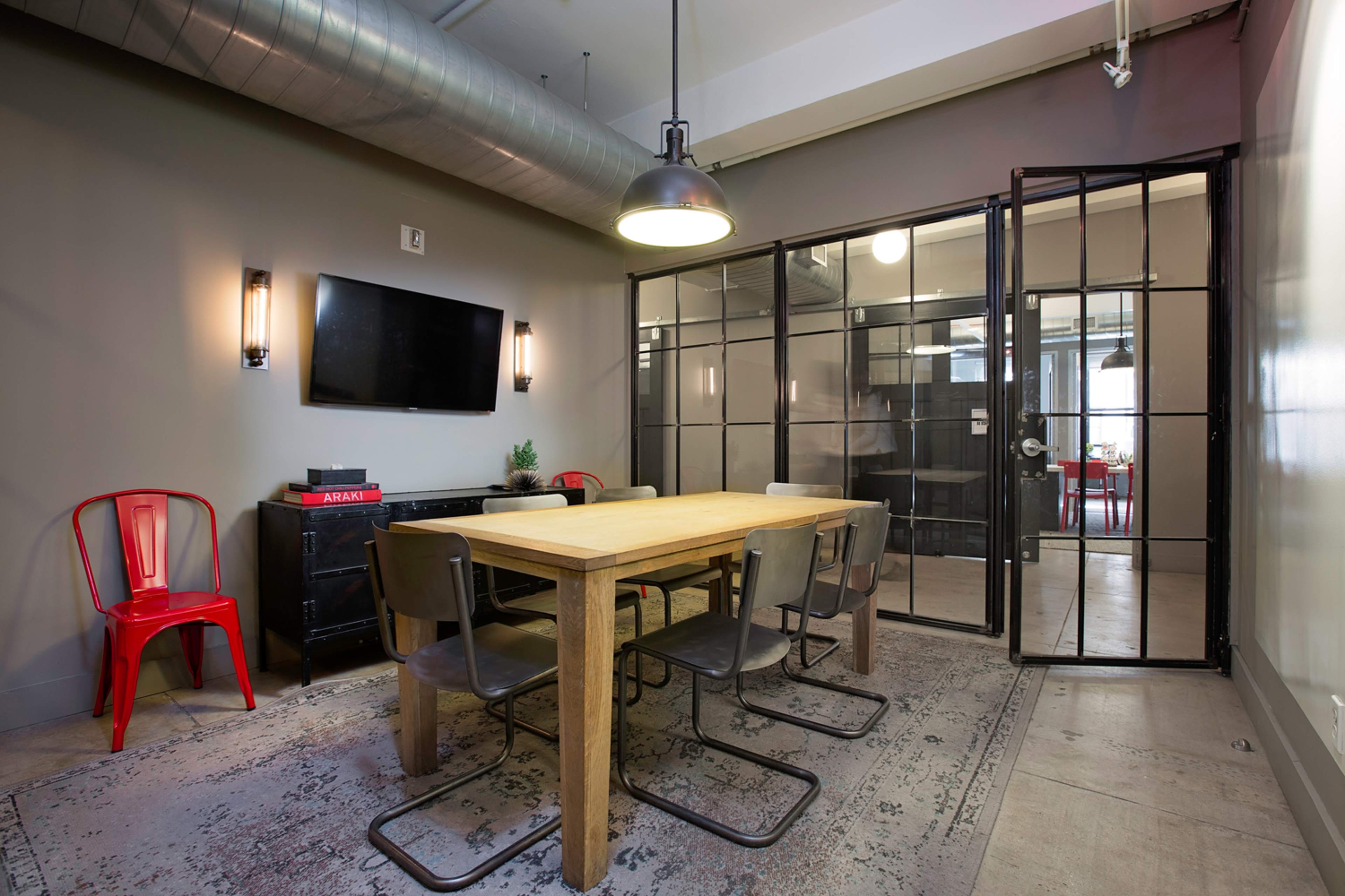 A conference room features a wooden table surrounded by metal chairs, with a large glass partition and a wall-mounted TV.