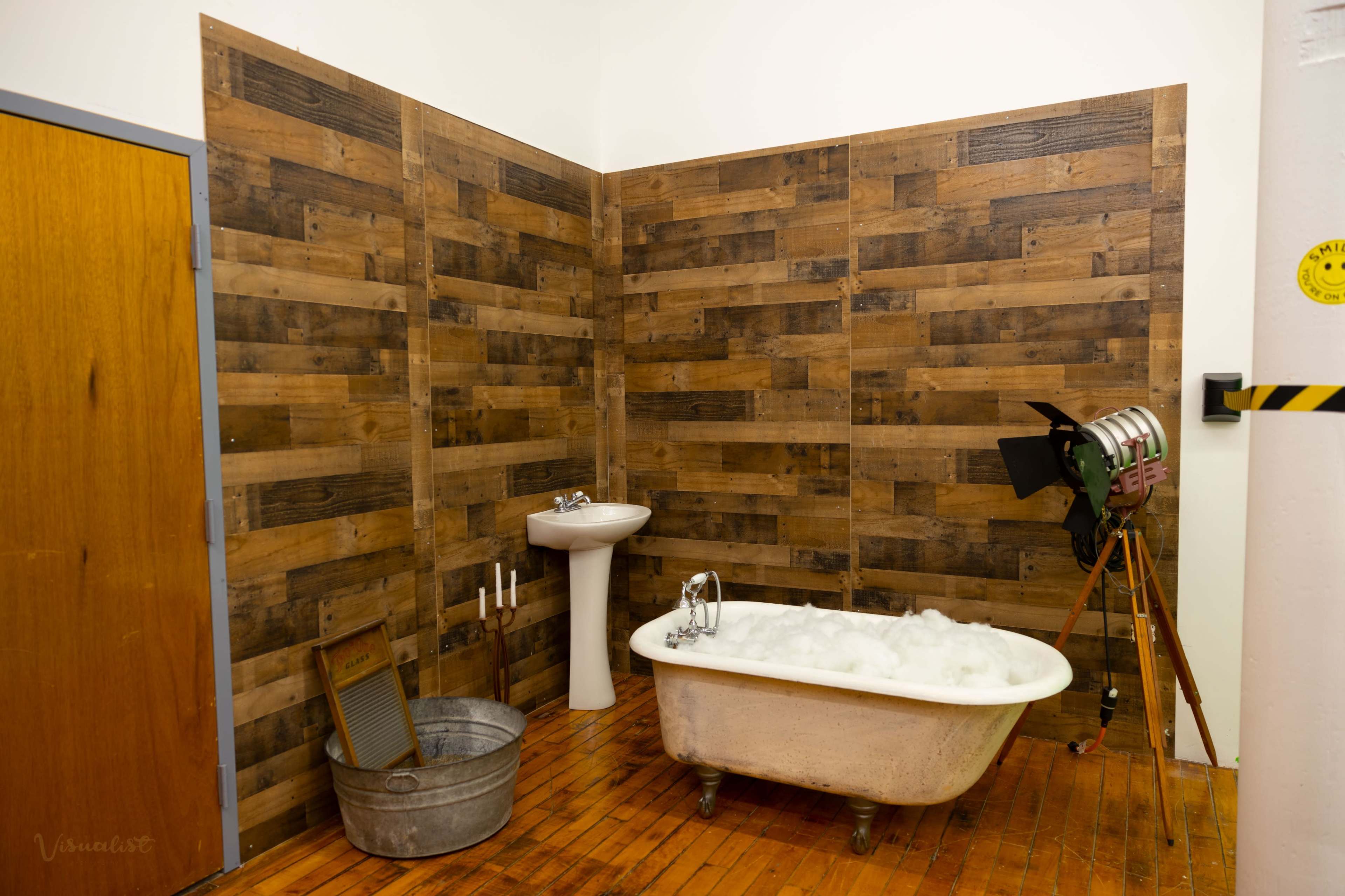 A bathroom setting with a freestanding bathtub filled with bubbles, a pedestal sink, a metal bucket, and a vintage tripod camera against a backdrop of wooden walls.