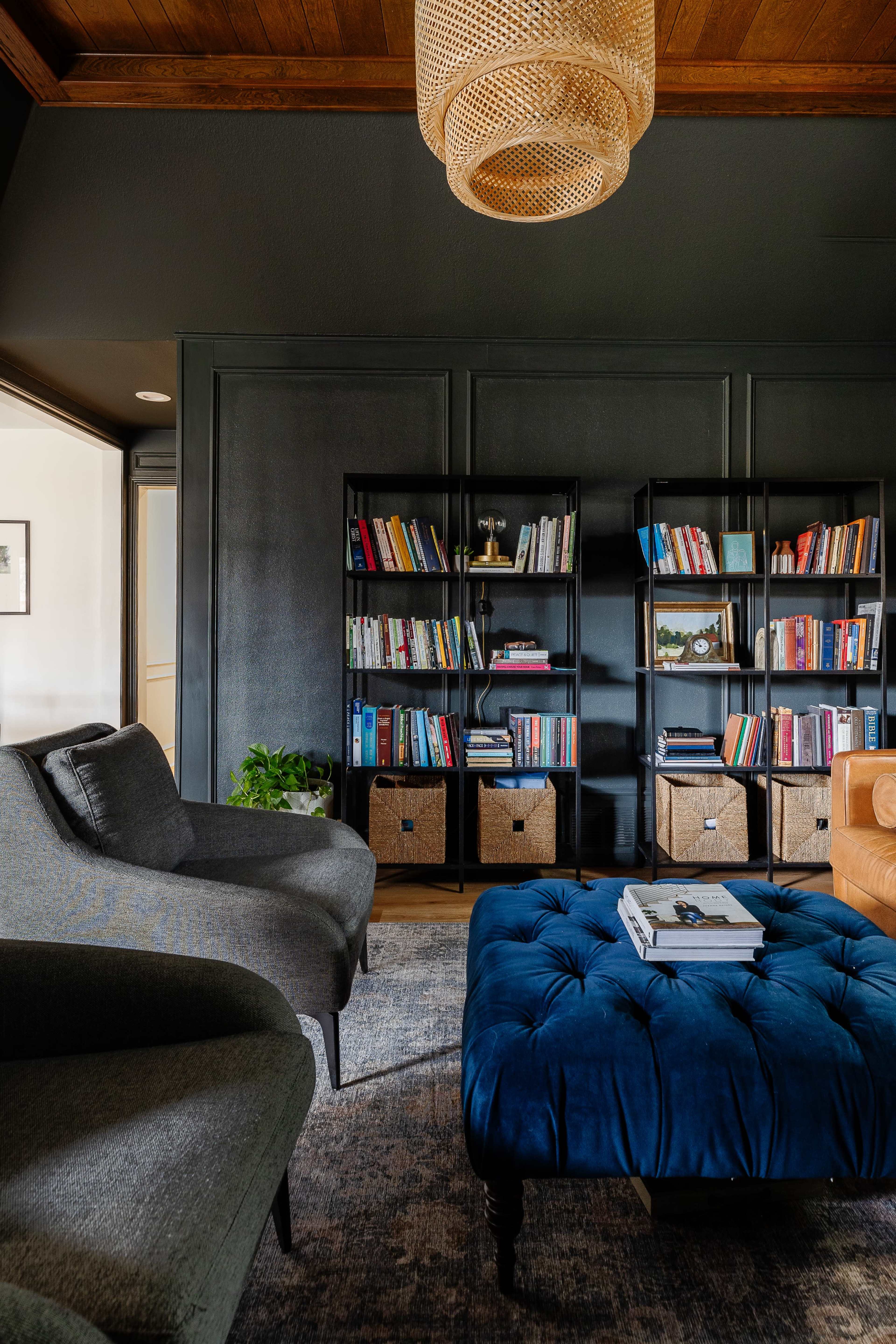 A cozy living room with dark walls, a velvet blue ottoman, two gray chairs, a bookshelf filled with books and decorative items, and a woven light fixture overhead.