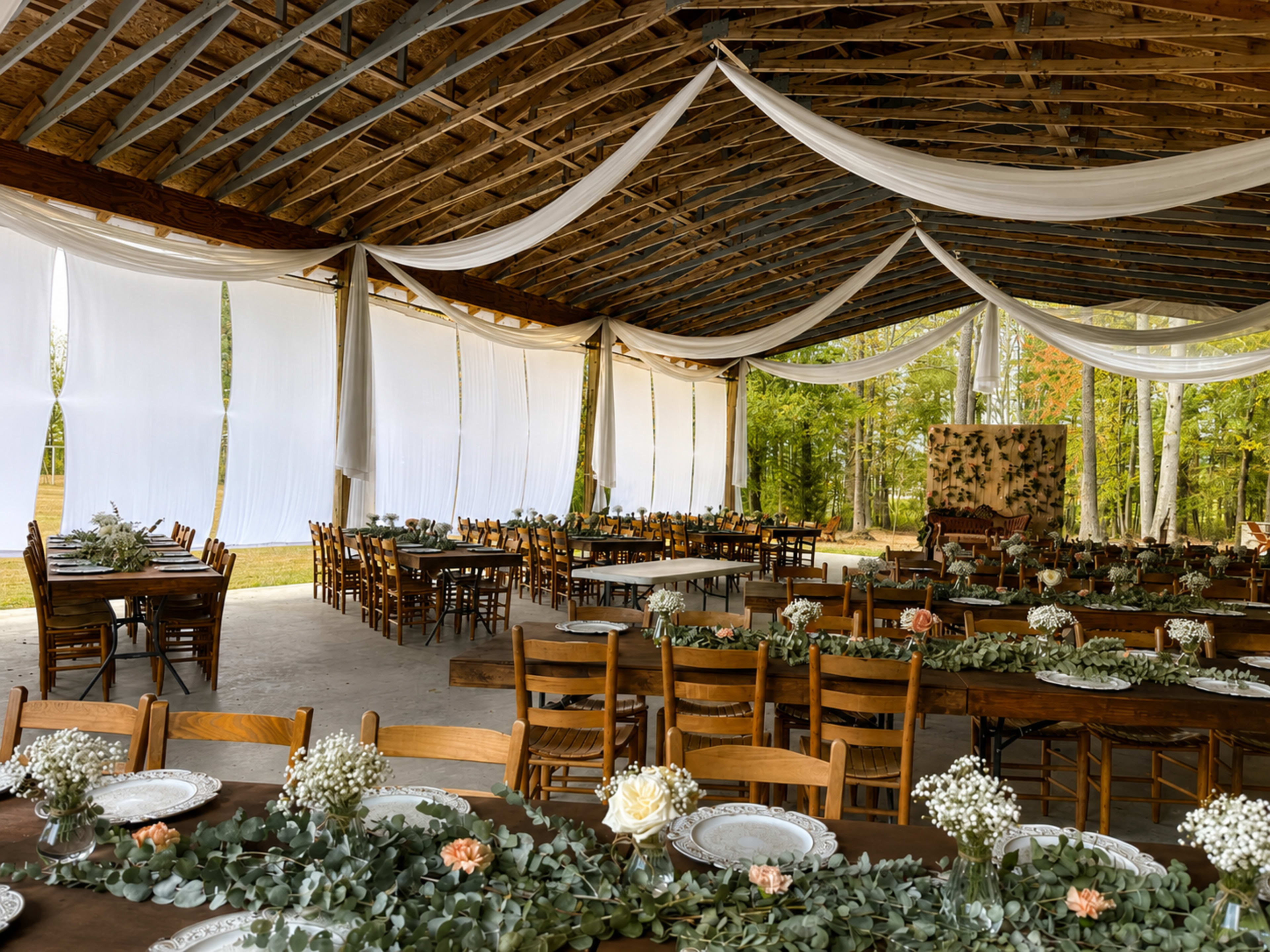 The image shows a rustic indoor venue decorated with draped fabric and wooden tables set for a reception, featuring floral centerpieces and wooden chairs.