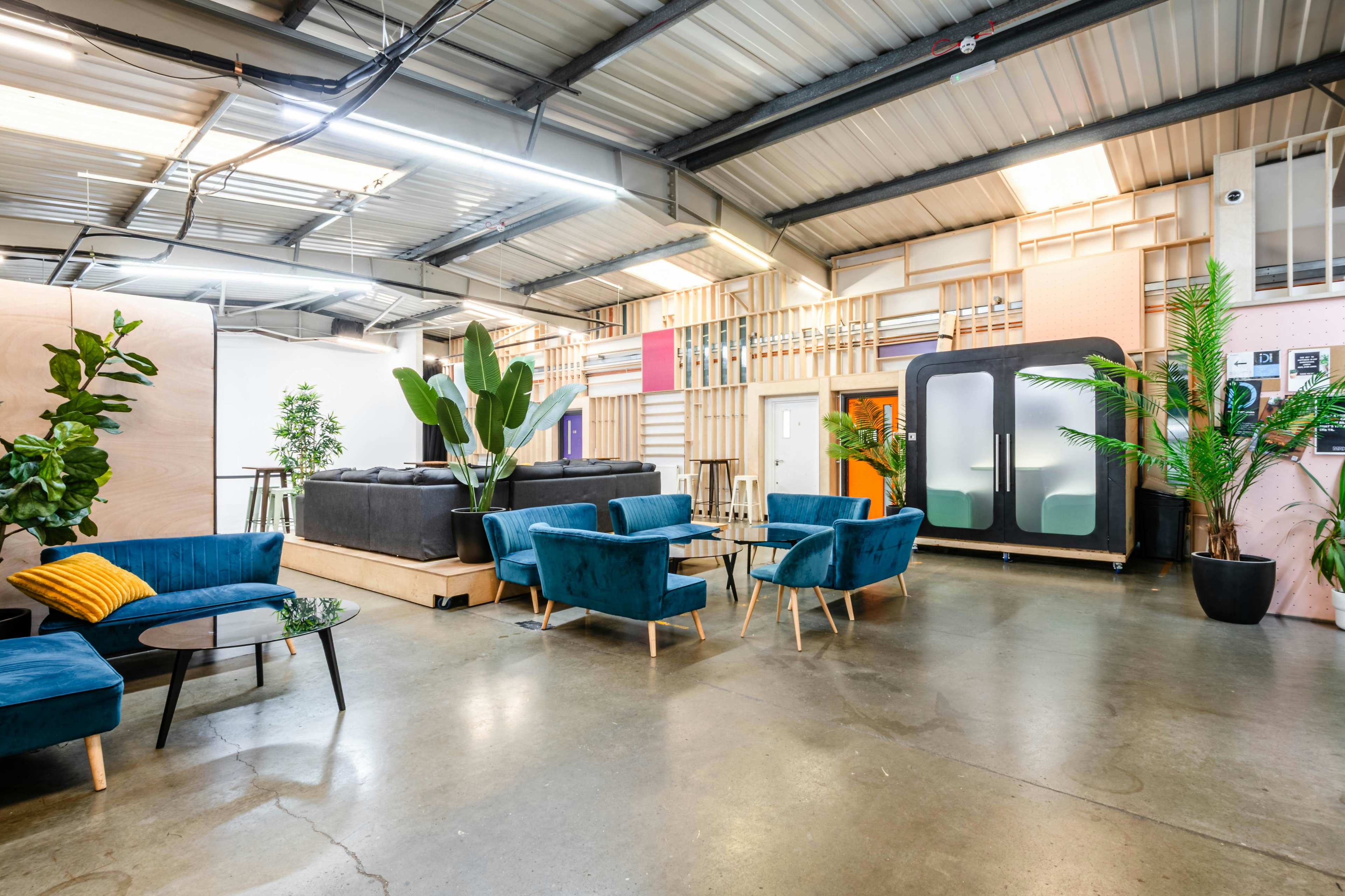 The image shows a modern workspace with a seating area featuring blue chairs, a black sofa, and green plants alongside a modular design in the background.